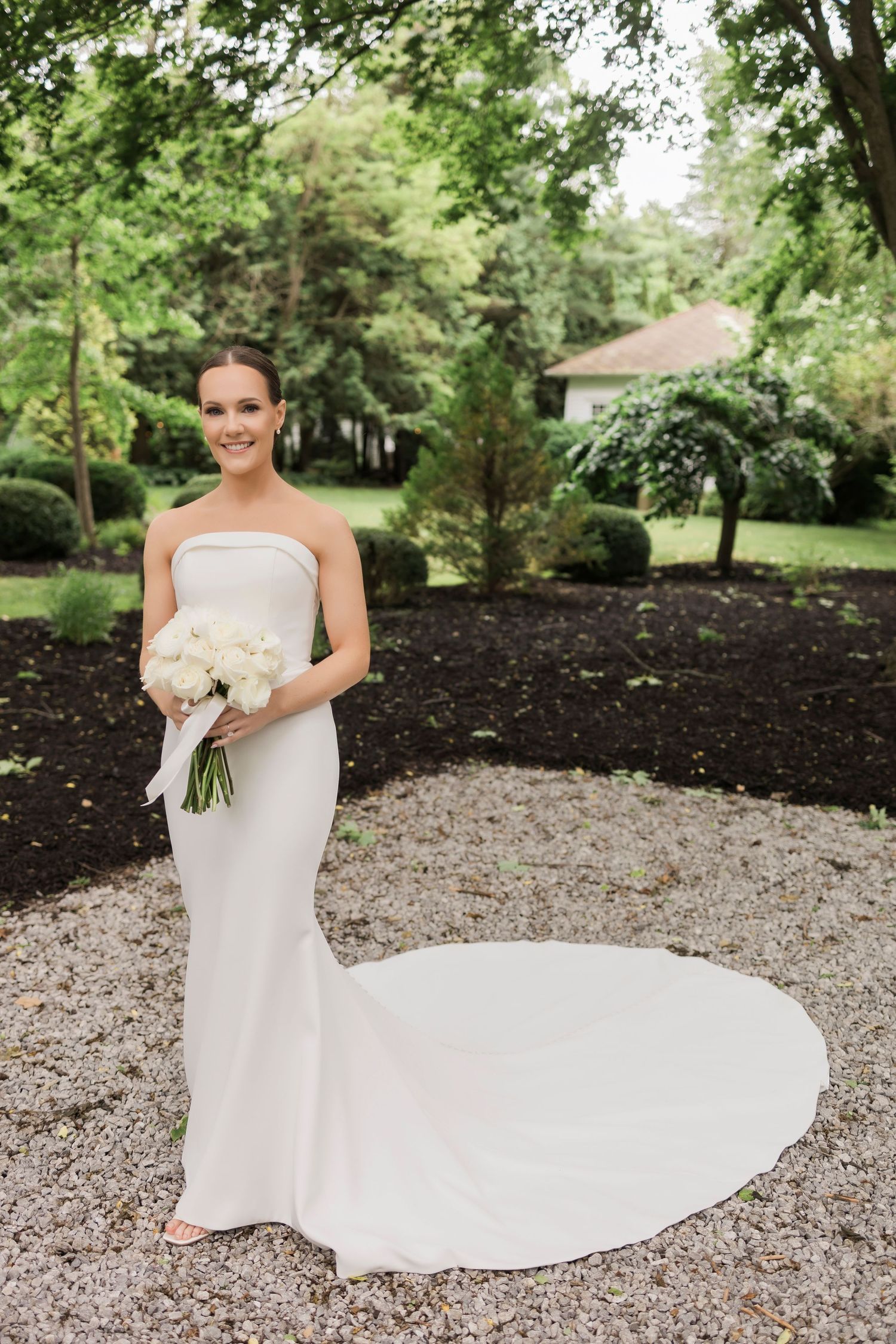 Elegant strapless white wedding dress with long train on garden path surrounded by lush greenery.