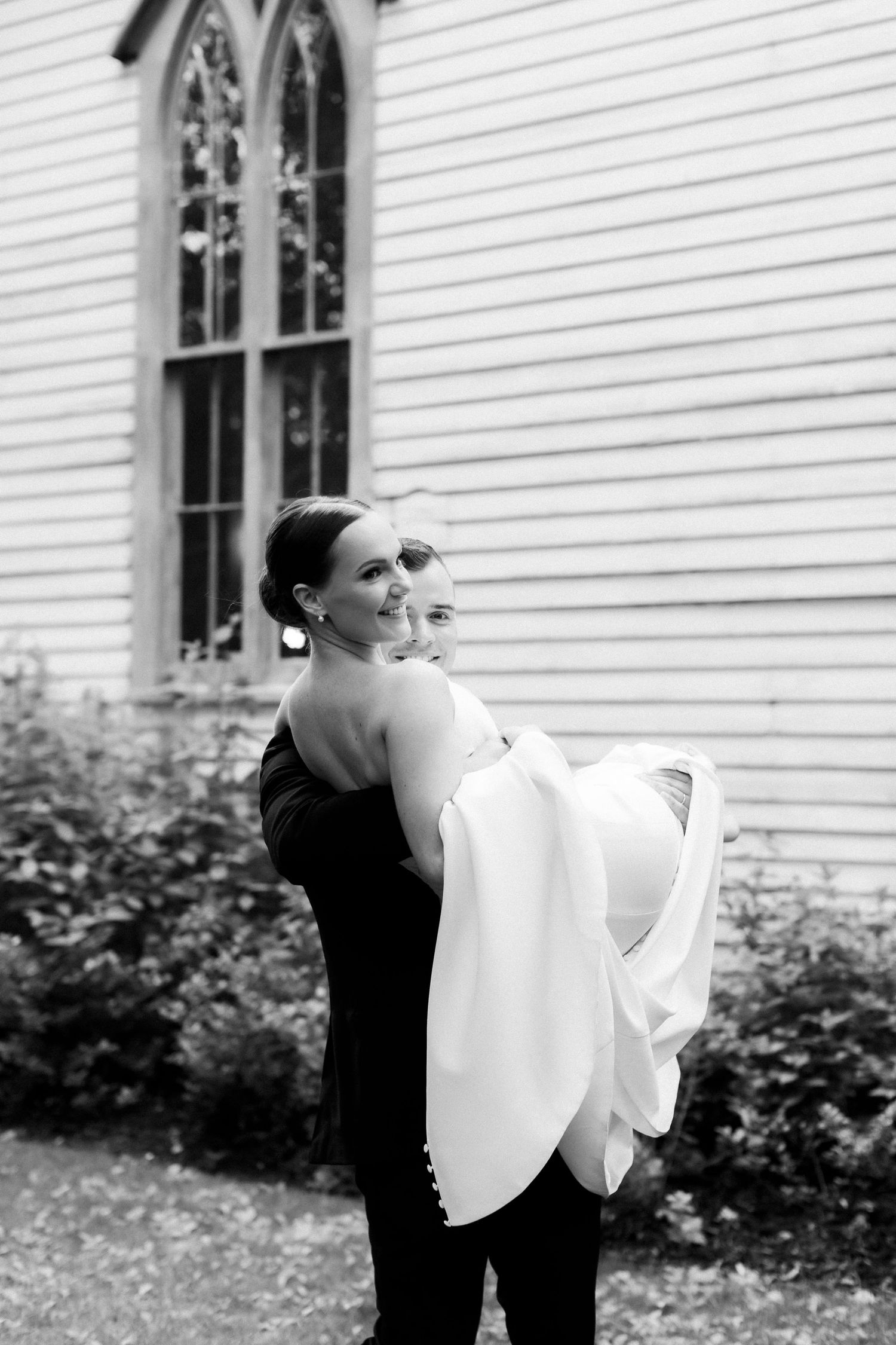 A black and white photo of a couple embracing in front of a white church with Gothic style windows.