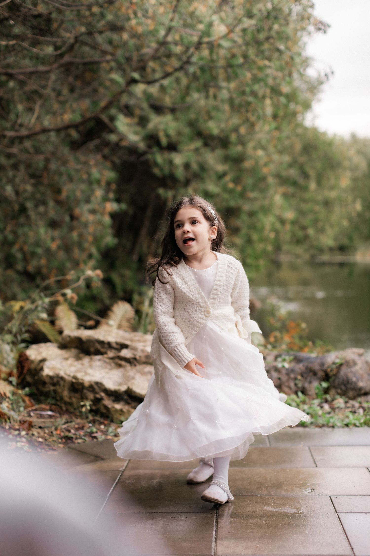 A young girl in a white lacy dress and cardigan twirls on stone pavement near a pond.