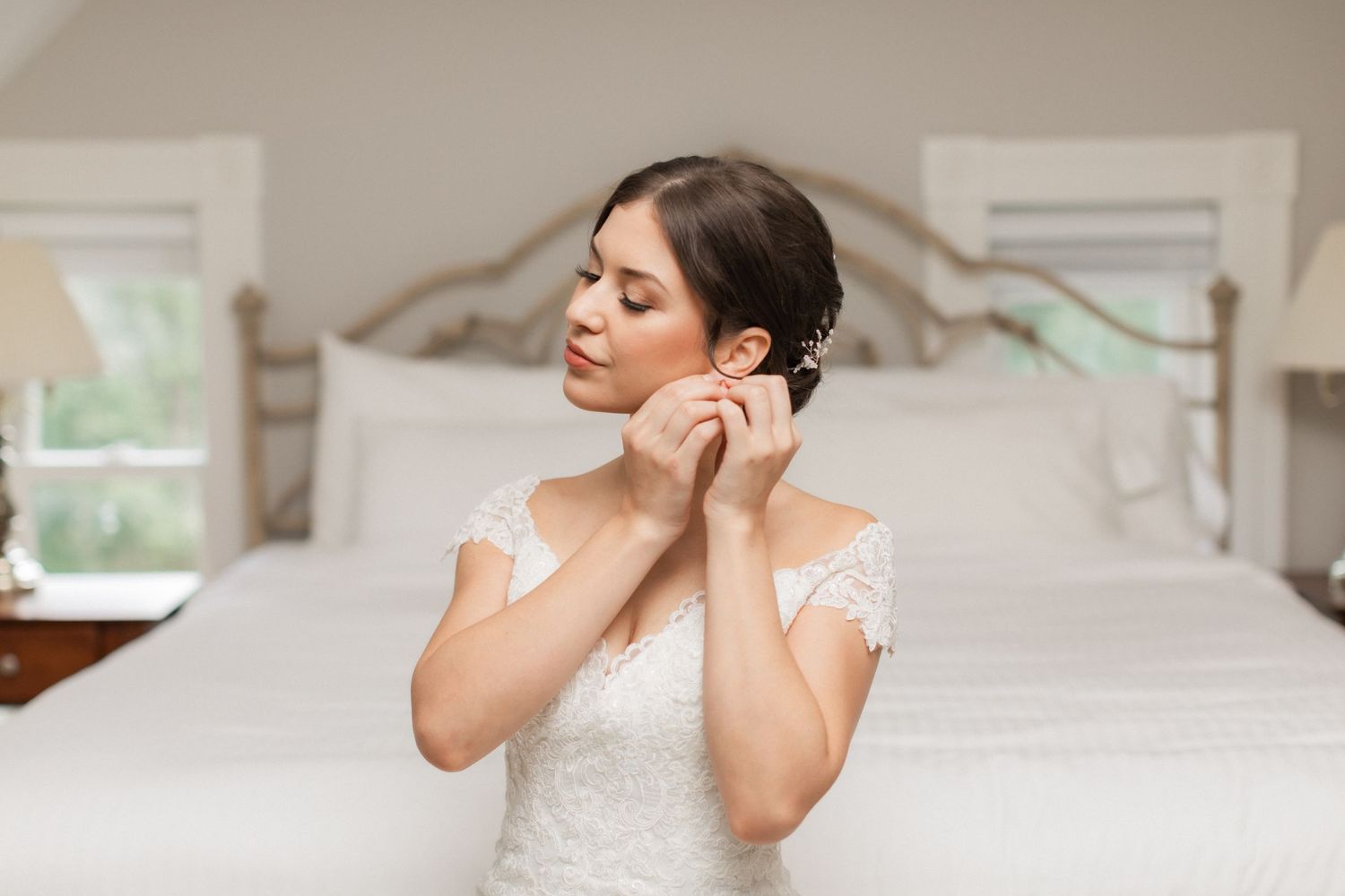 Bride putting on earrings while getting ready in elegant bedroom setting.