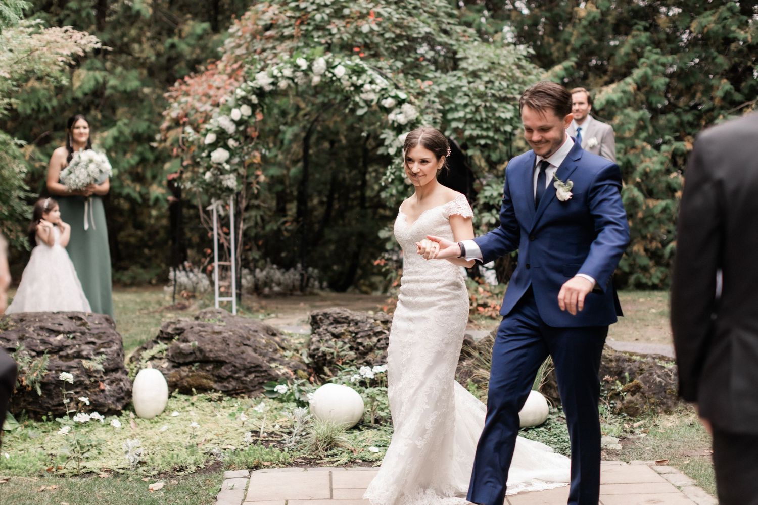 Wedding couple elegantly exit their ceremony through a stone pathway in a lush garden setting.