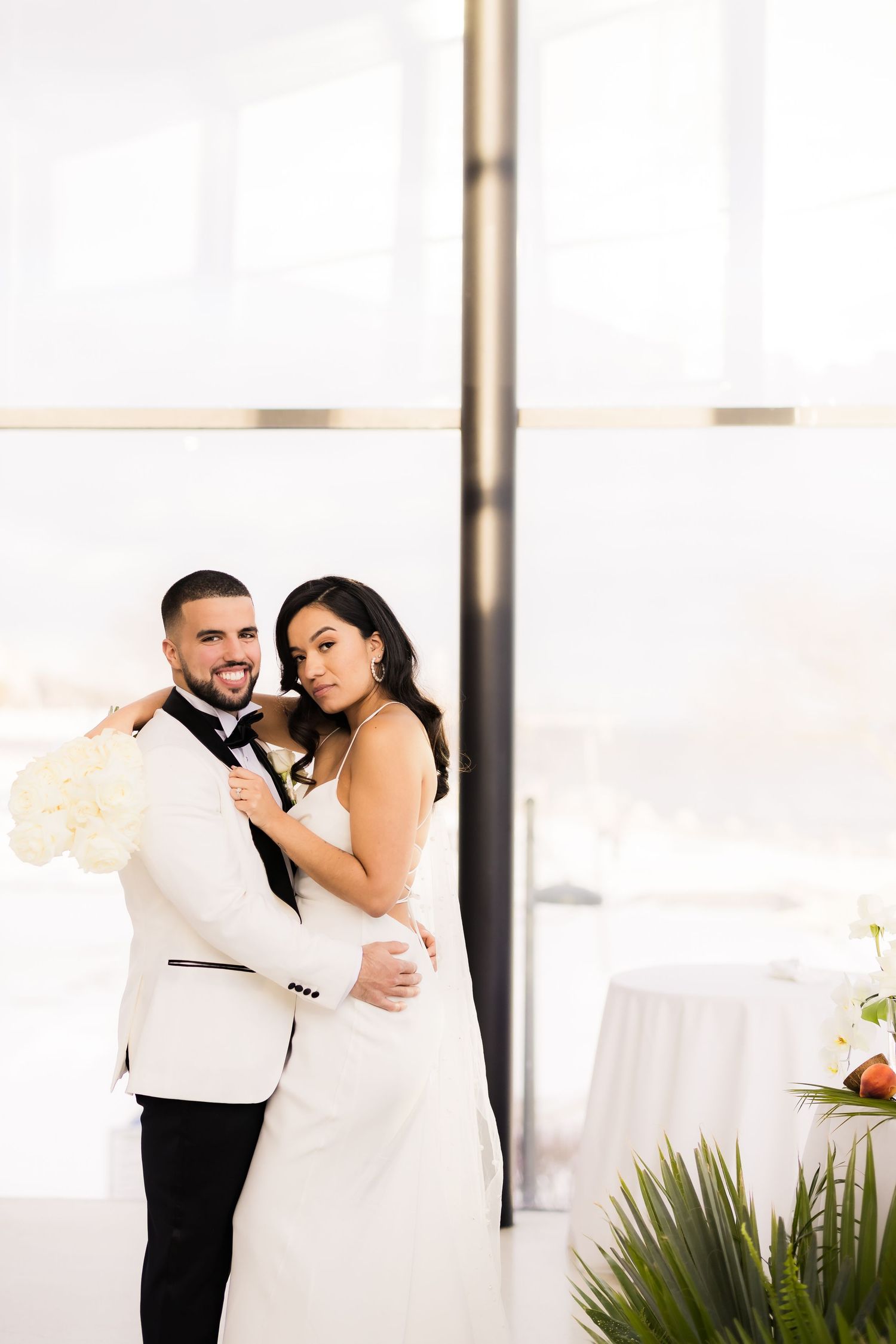 A wedding couple poses elegantly by a modern pillar with palm decor and white flowers.