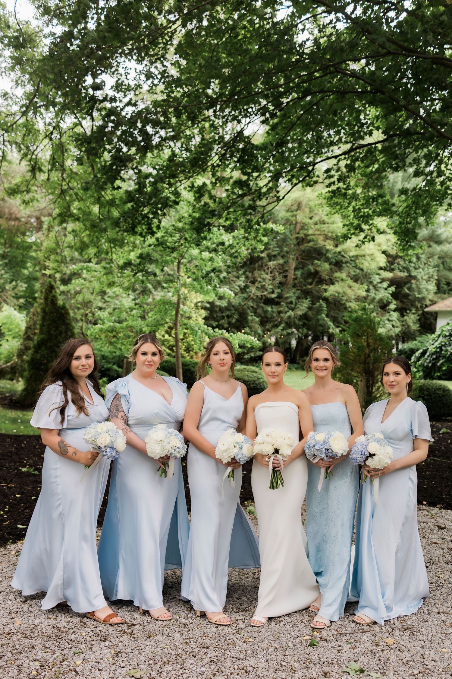 Wedding party in long pale blue dresses and white gown stand together on gravel path surrounded by lush greenery.