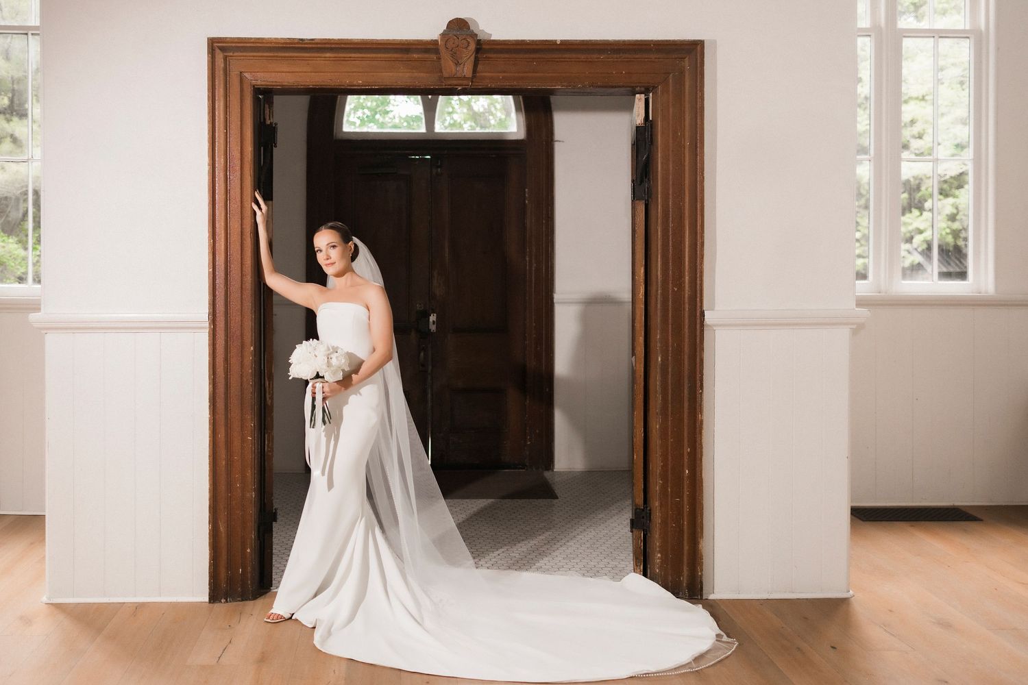 A bride in a flowing white satin dress with a long train stands elegantly in a doorway with natural lighting.