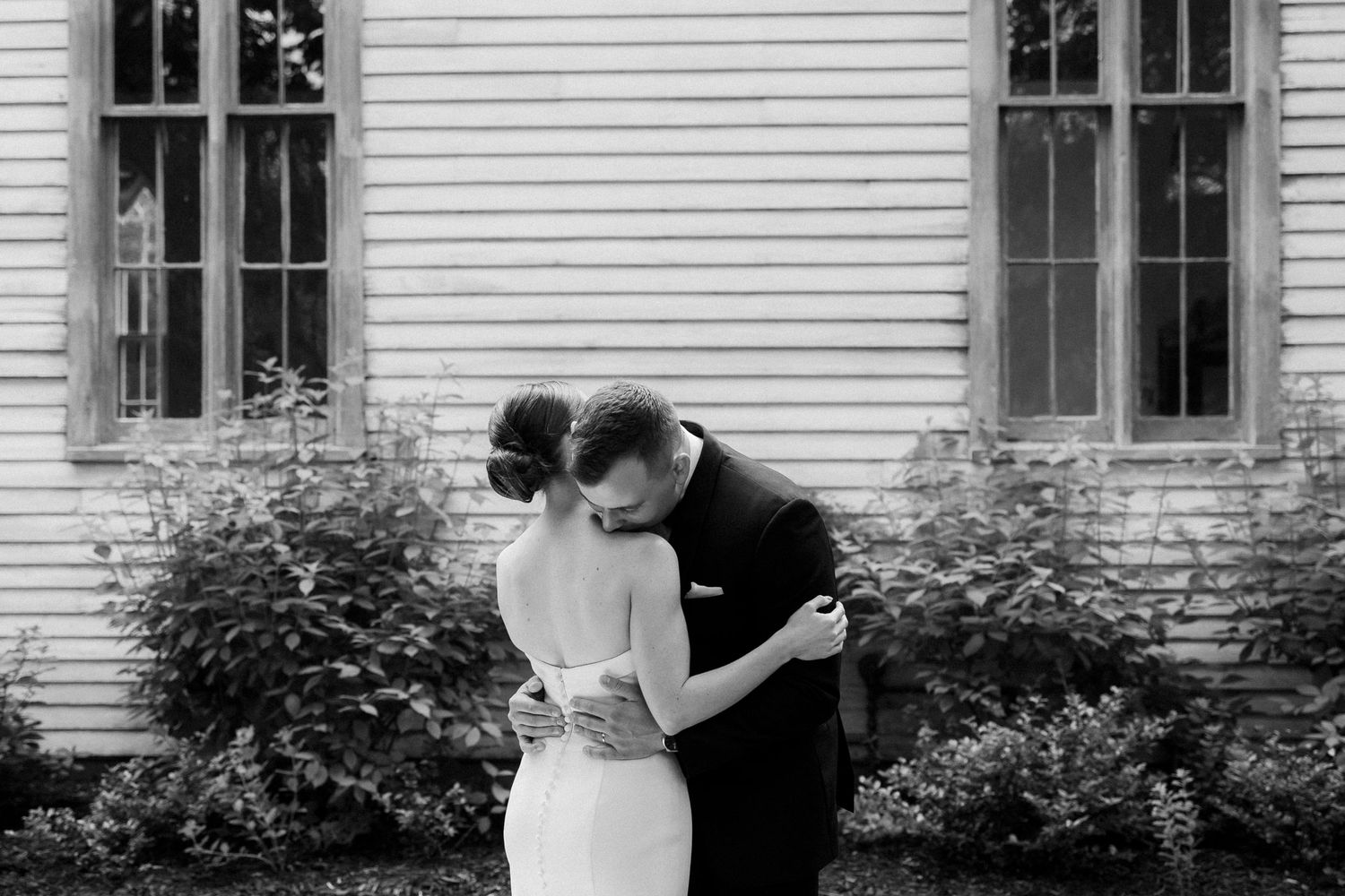 A tender moment shared between a couple in formal wedding attire embracing on a porch in black and white.
