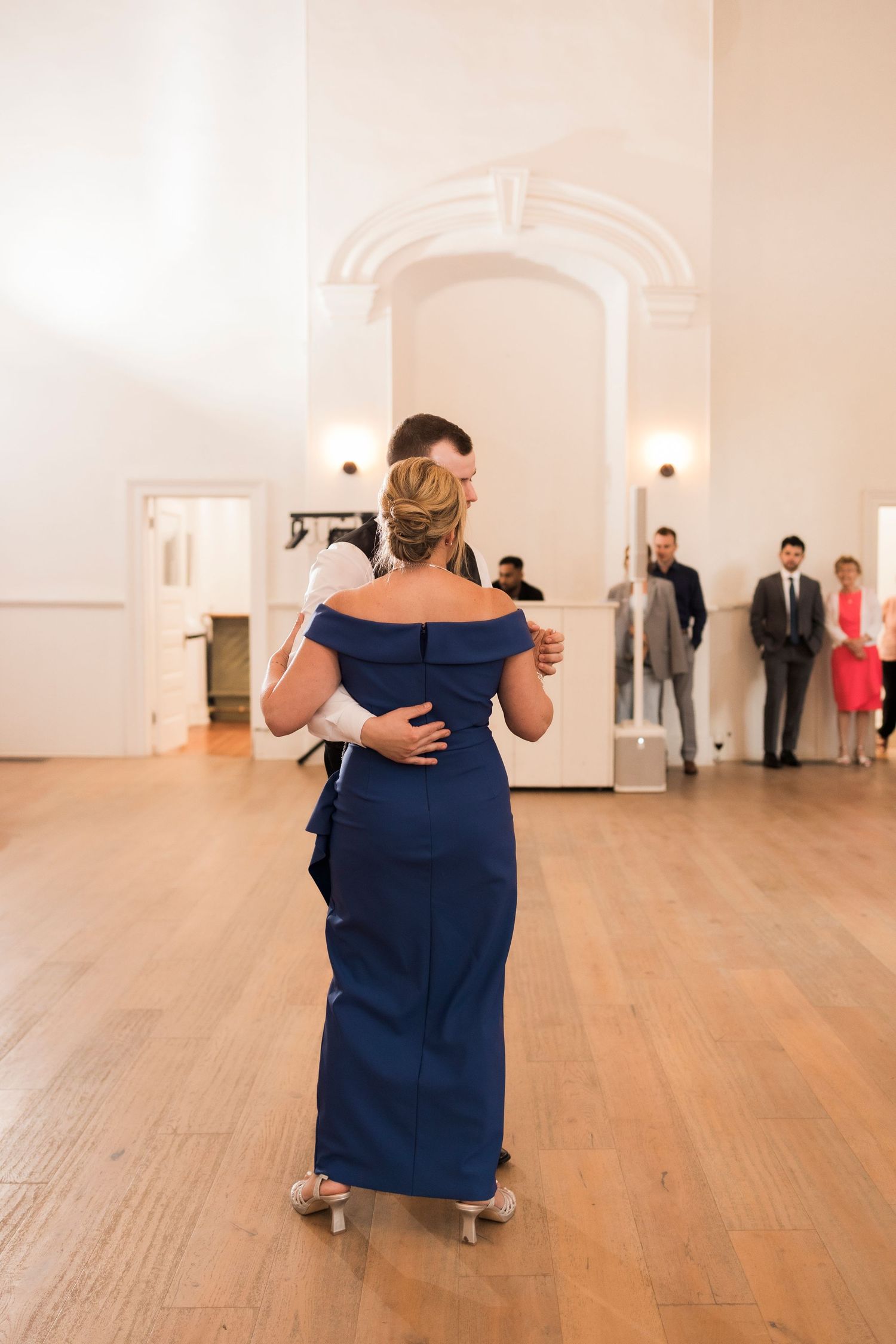 An elegant off-shoulder navy blue gown is displayed from behind on the wooden floor of a bright, arched venue.