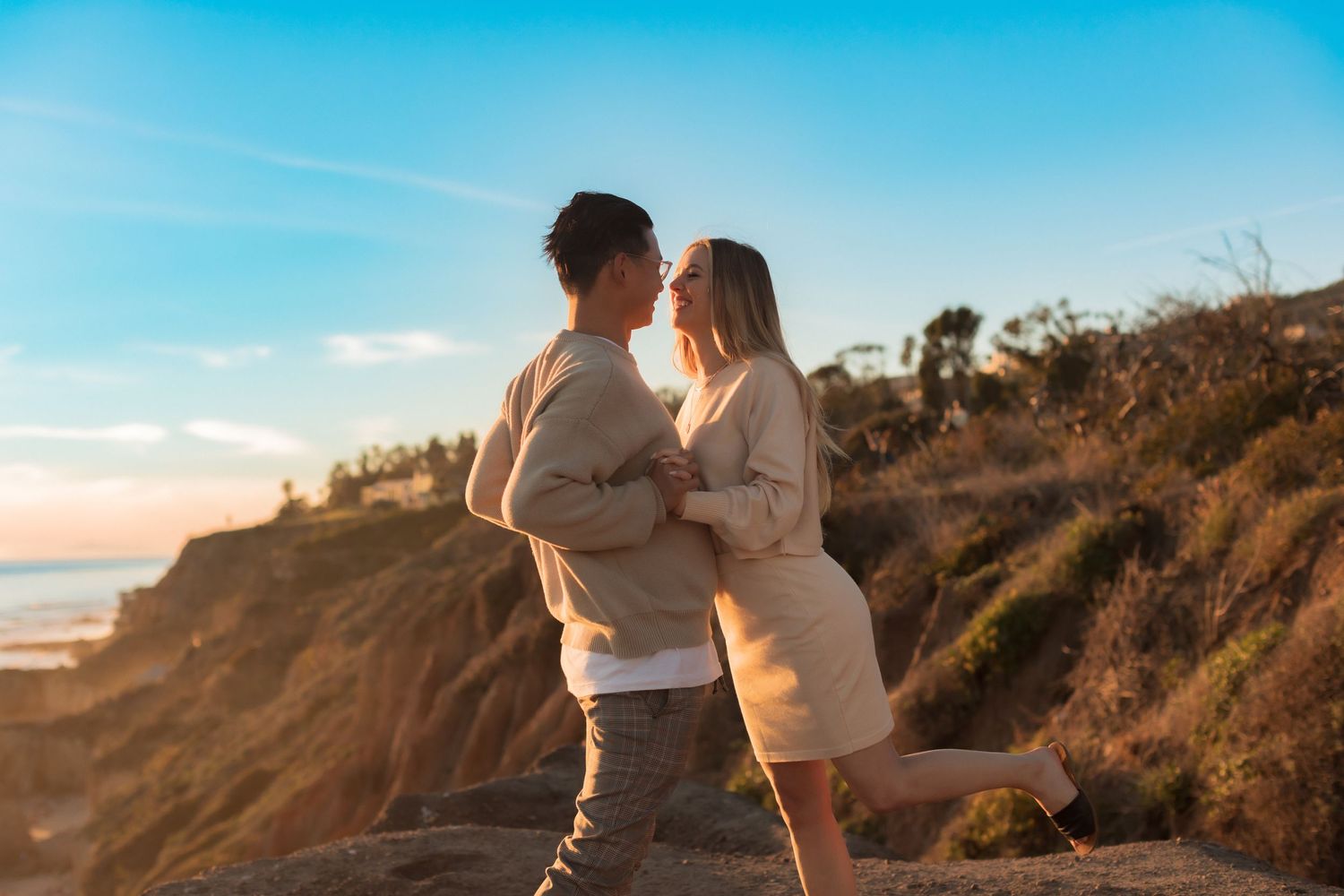 Couple in light-colored outfits embrace on dramatic clifftop overlooking ocean waves.