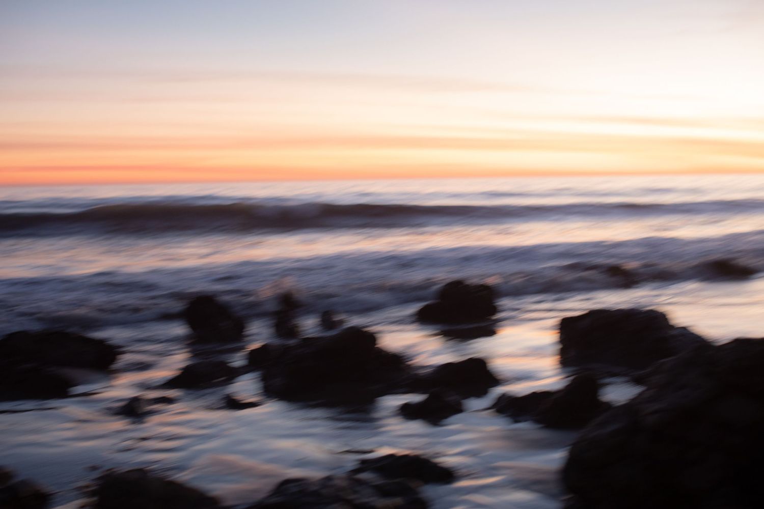Long exposure seascape at sunset with blurred waves crashing over dark rocks.