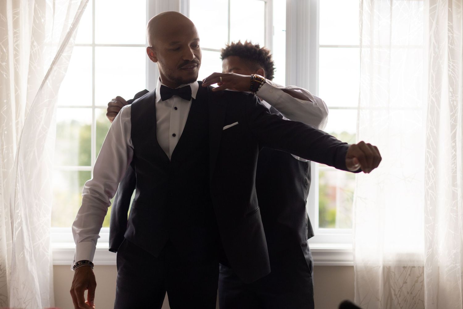 Two figures in formal attire assist each other with wedding preparations.