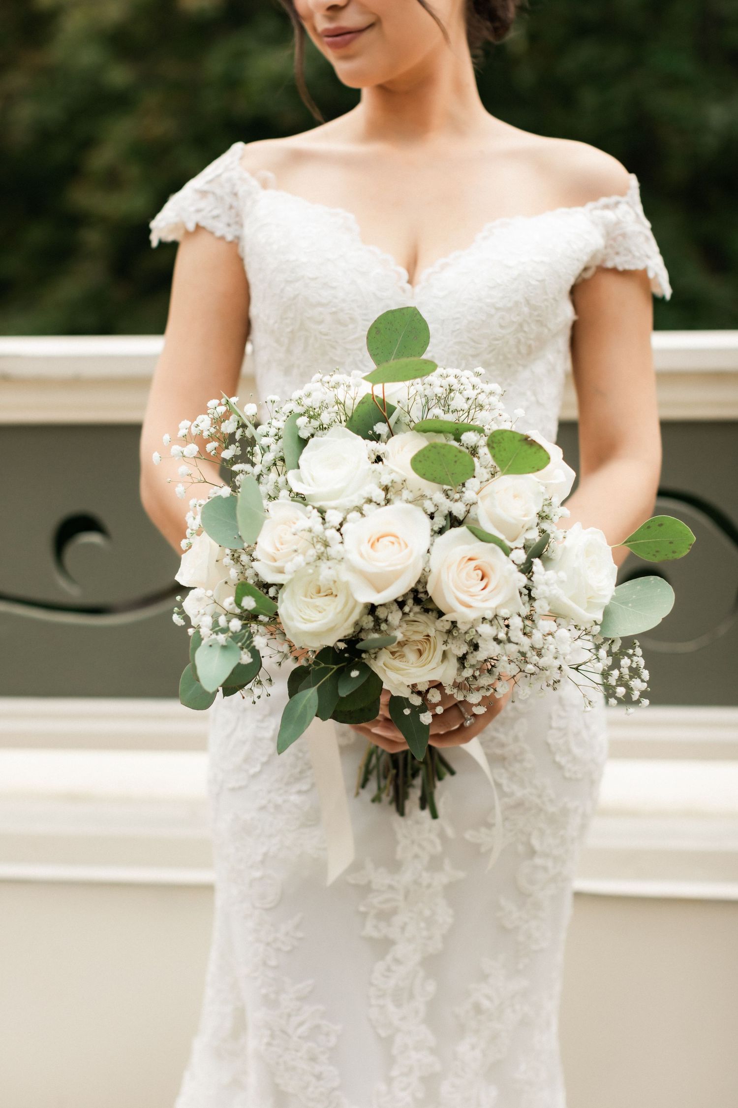 Close-up of white and green bridal bouquet with roses and eucalyptus against lace wedding dress.