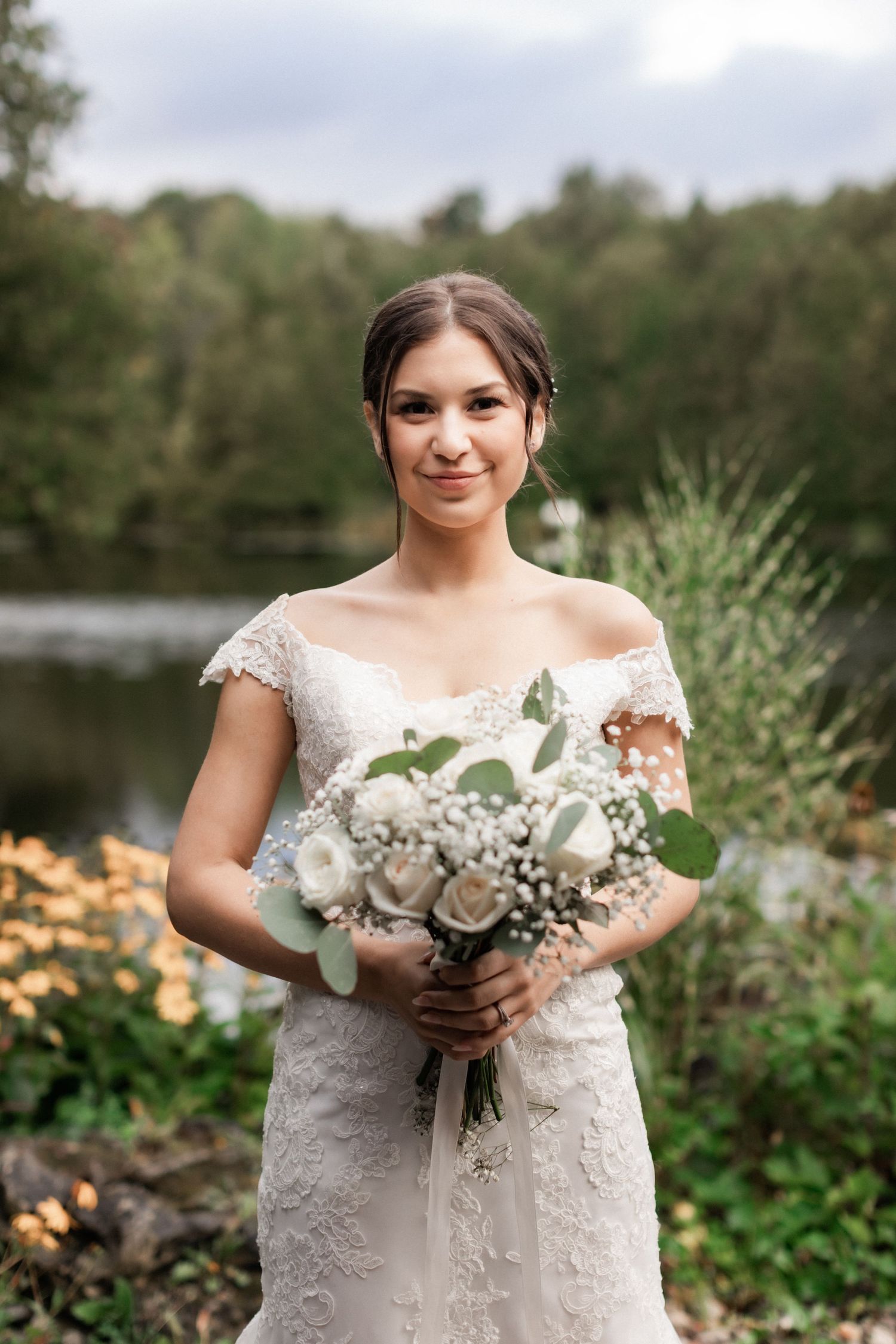 An elegant bridal portrait featuring a lace wedding dress and white bouquet by a lake.