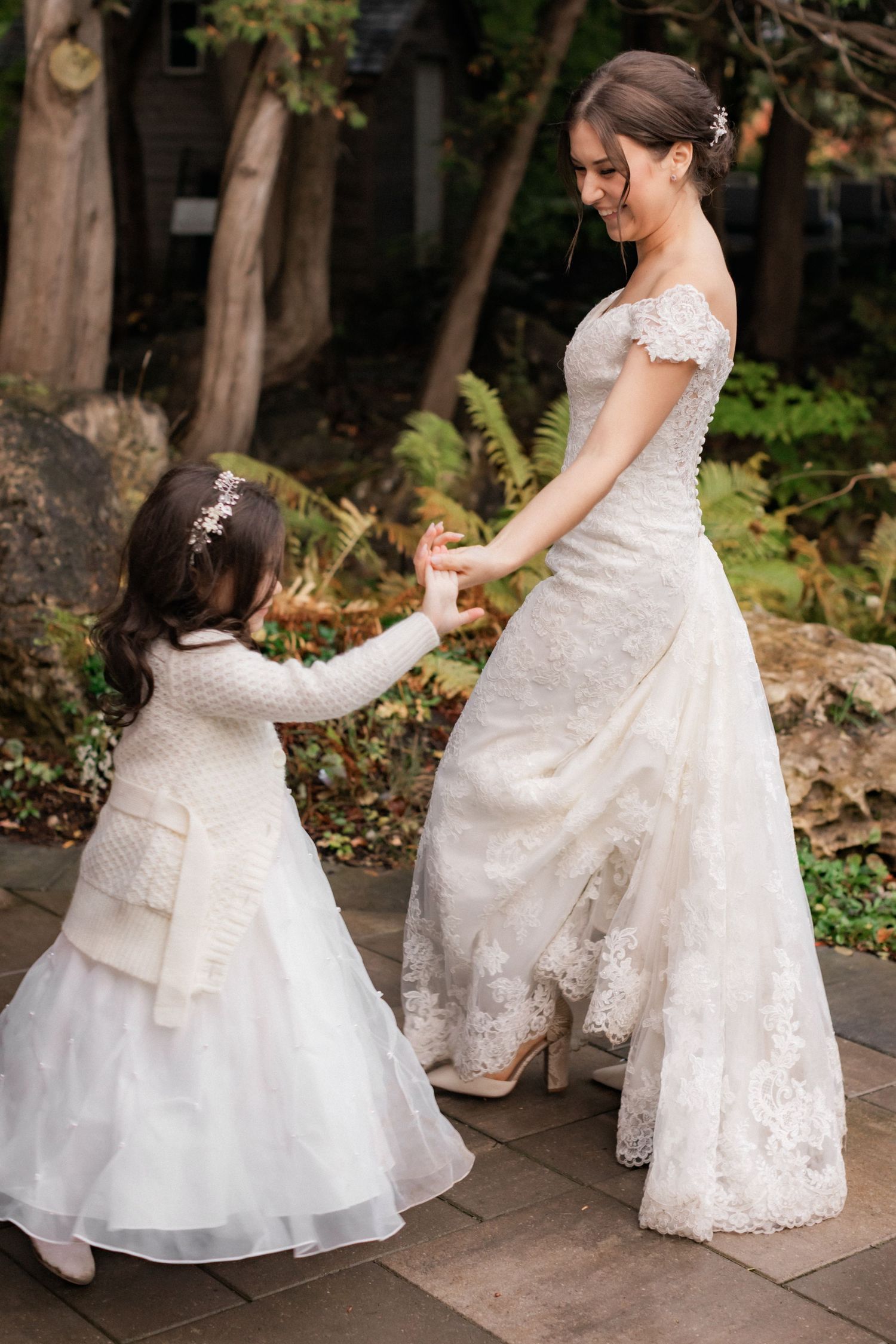 A touching moment between a bride and young flower girl in matching white lace dresses holding hands outdoors.