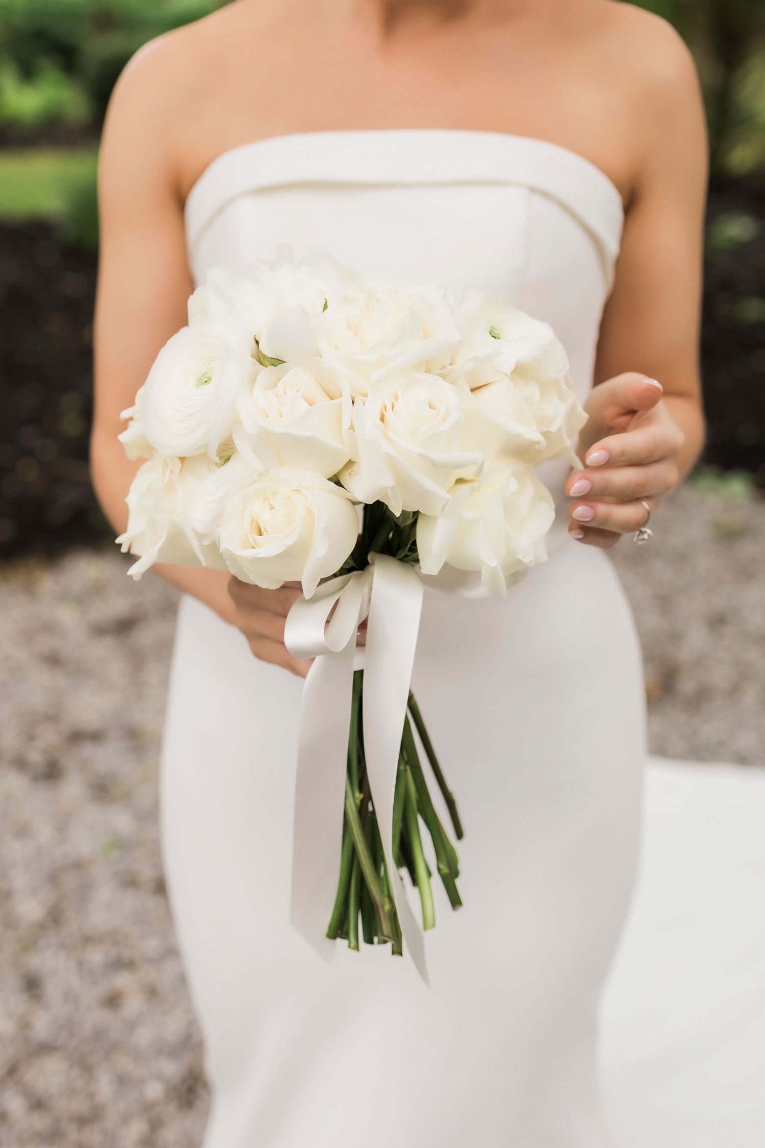 Elegant bride in strapless white gown shown with pristine white rose bouquet outdoors.