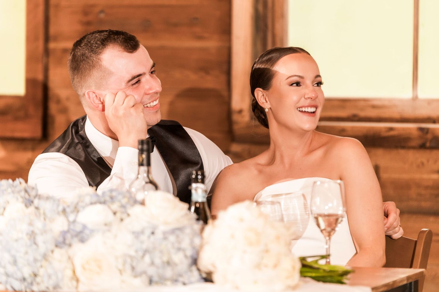 People seated at rustic wooden tables during a wedding reception with bright lighting.