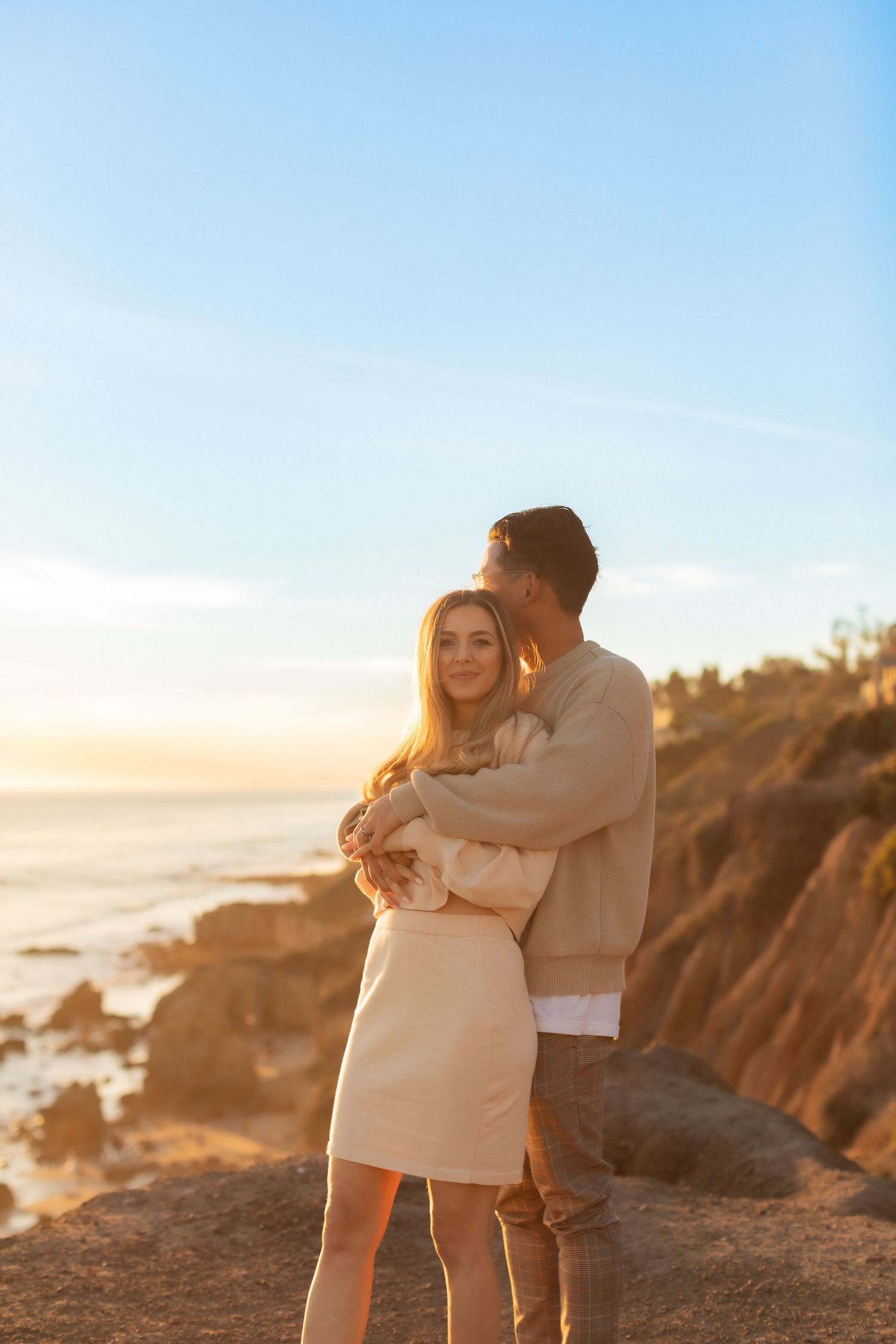 Couple in neutral tones sharing intimate embrace on rocky coastal overlook.