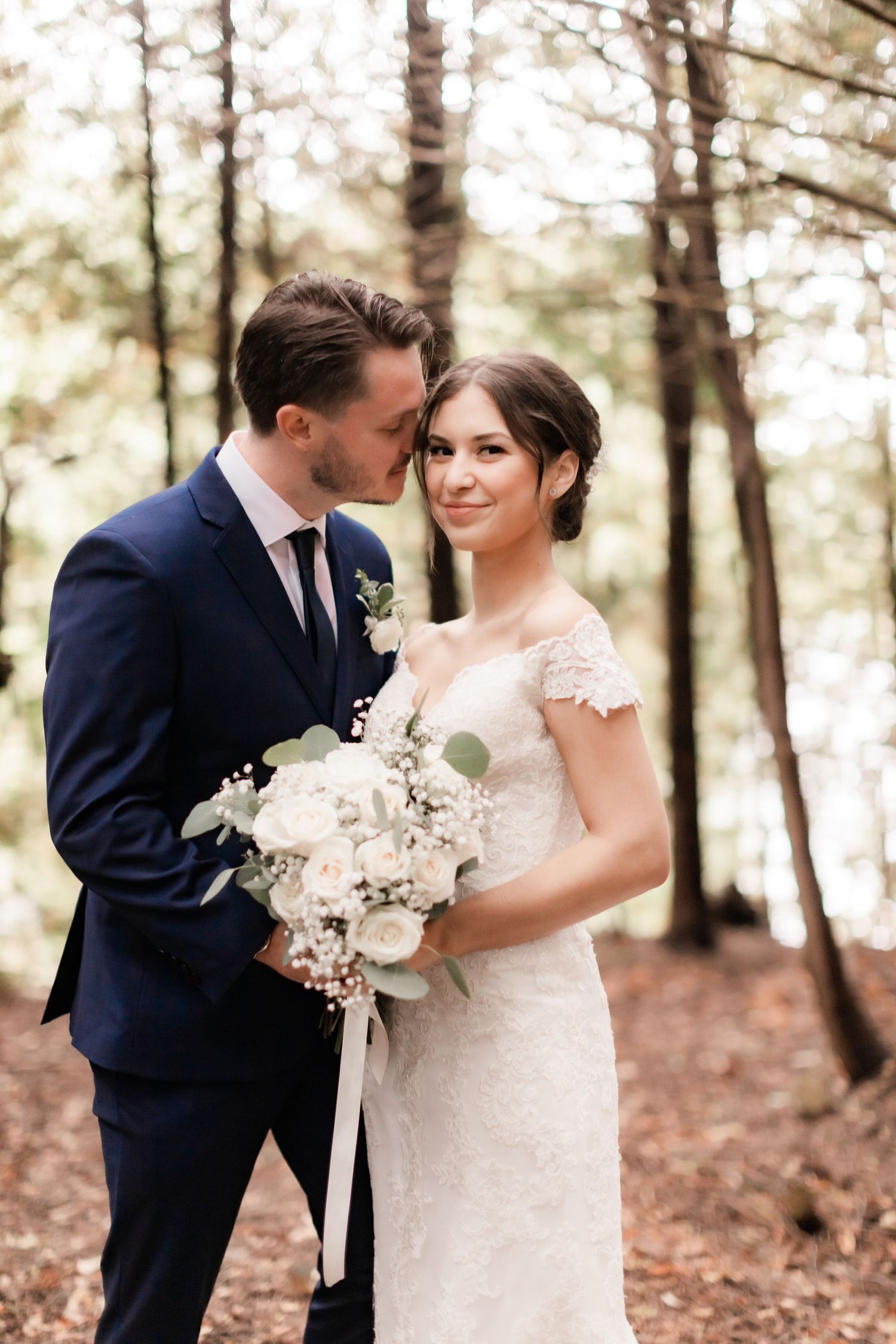 Intimate forest wedding portrait with couple holding white bouquet among pine trees.