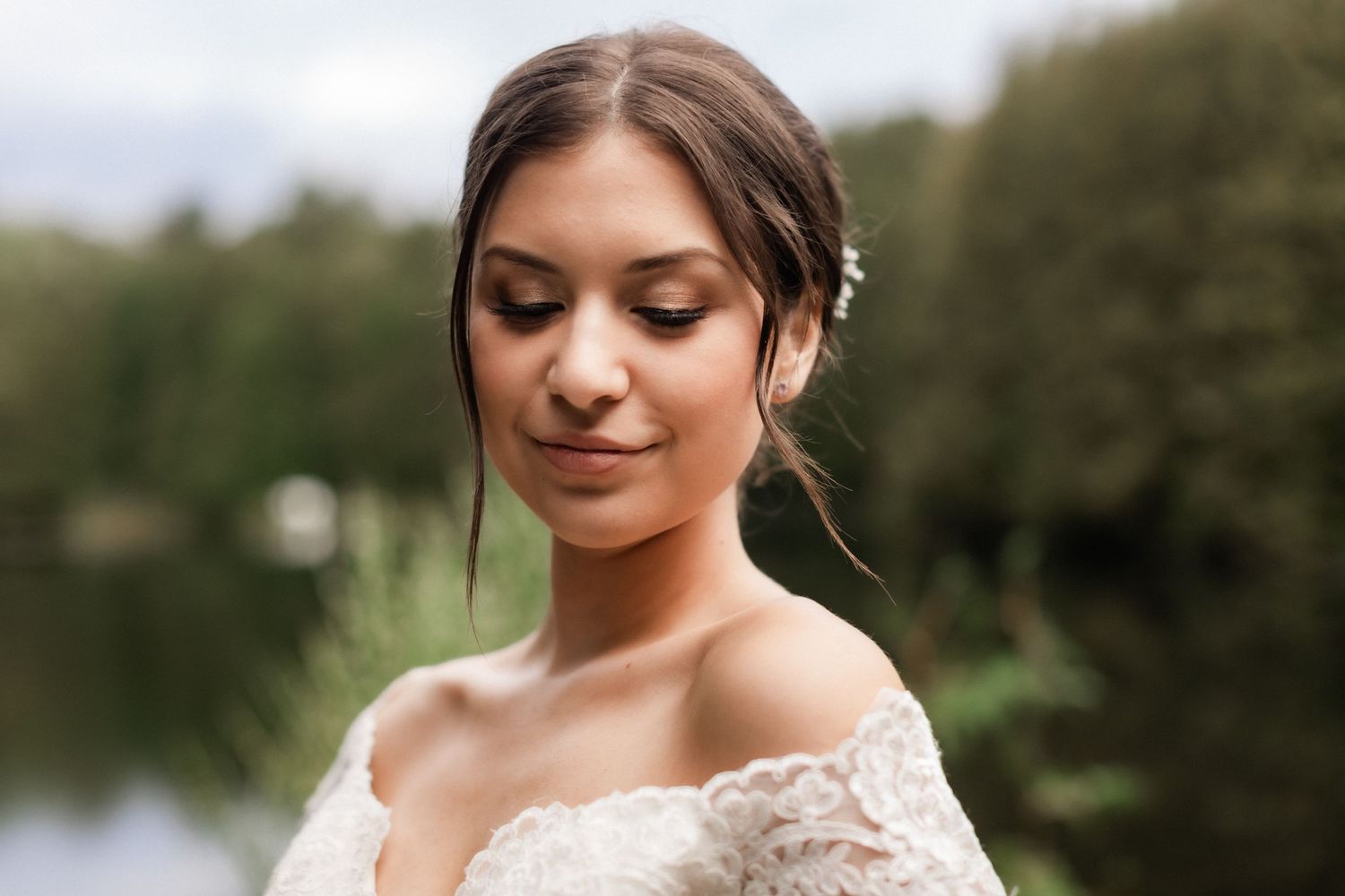 Close-up portrait of a bride in lace with natural makeup against a lake backdrop.