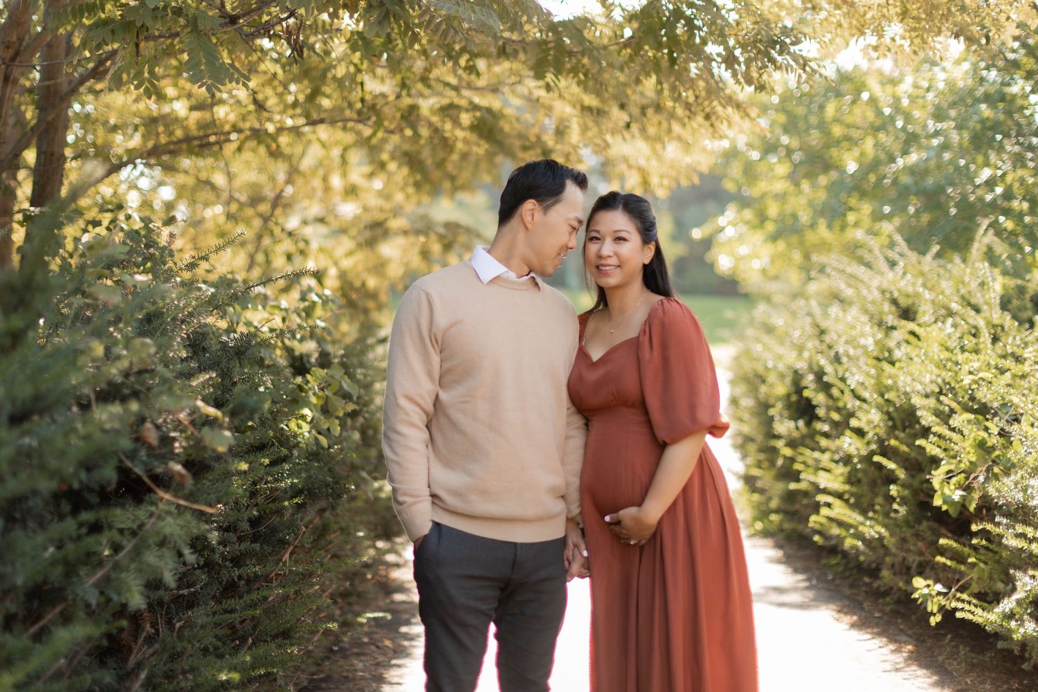 Couple poses on garden path surrounded by greenery during sunset maternity photography.