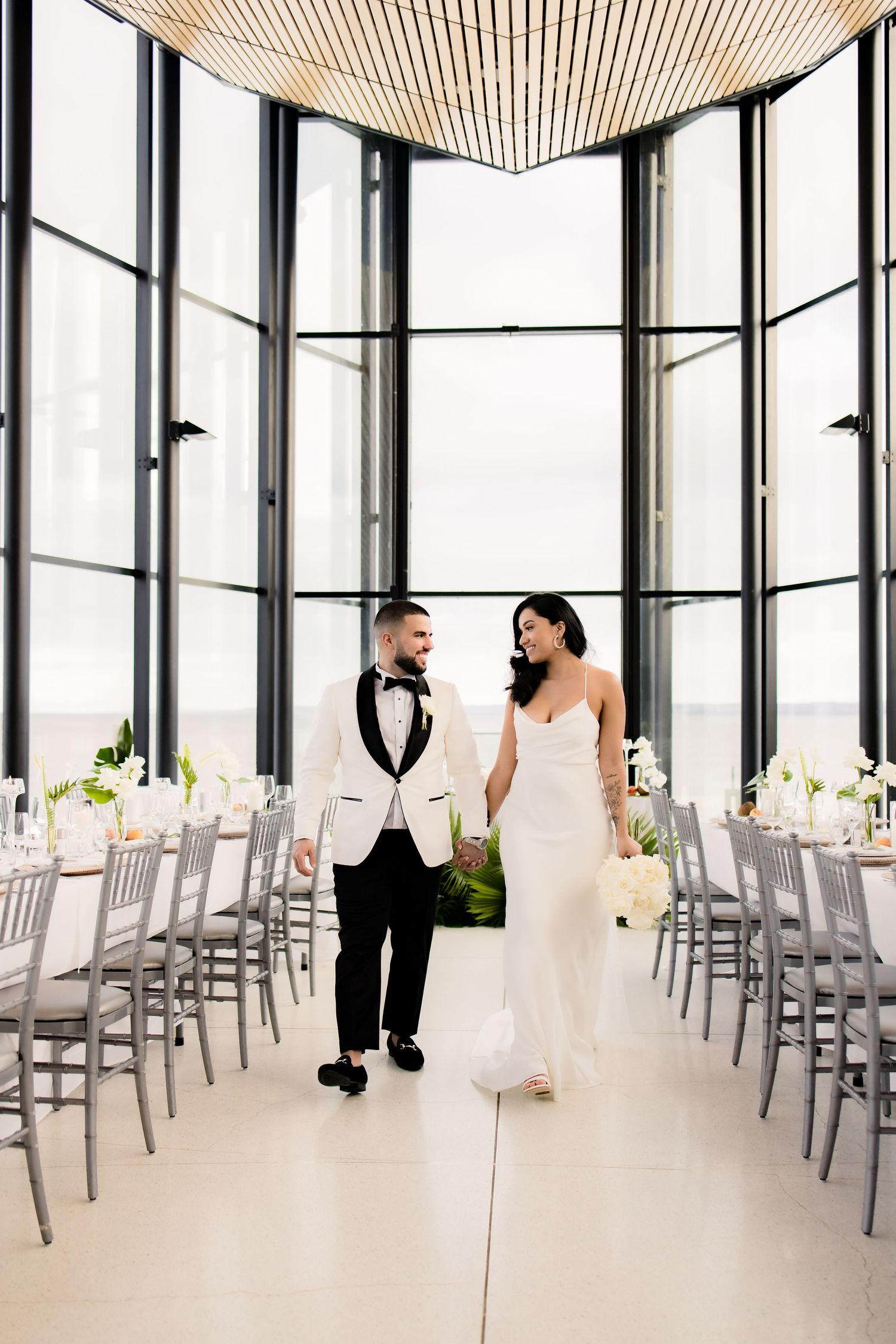 Wedding couple walking through modern venue with glass walls and elegant table settings.