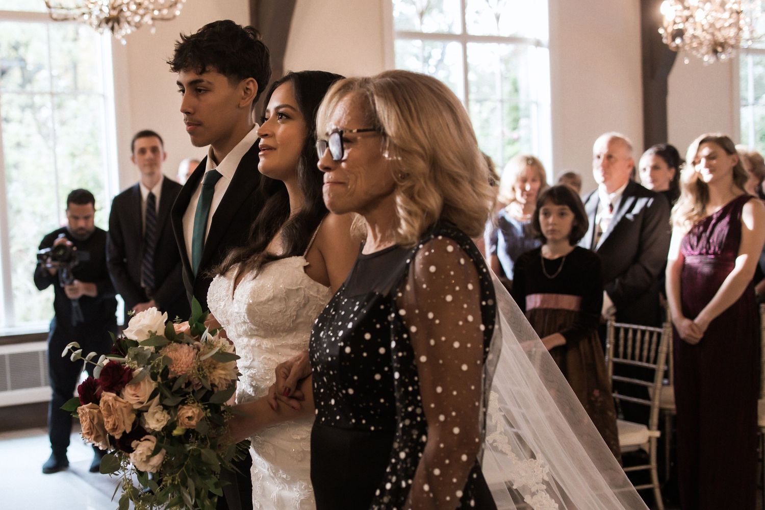 Couple sharing emotional moment during indoor wedding ceremony with sparkly-dressed guest looking on.