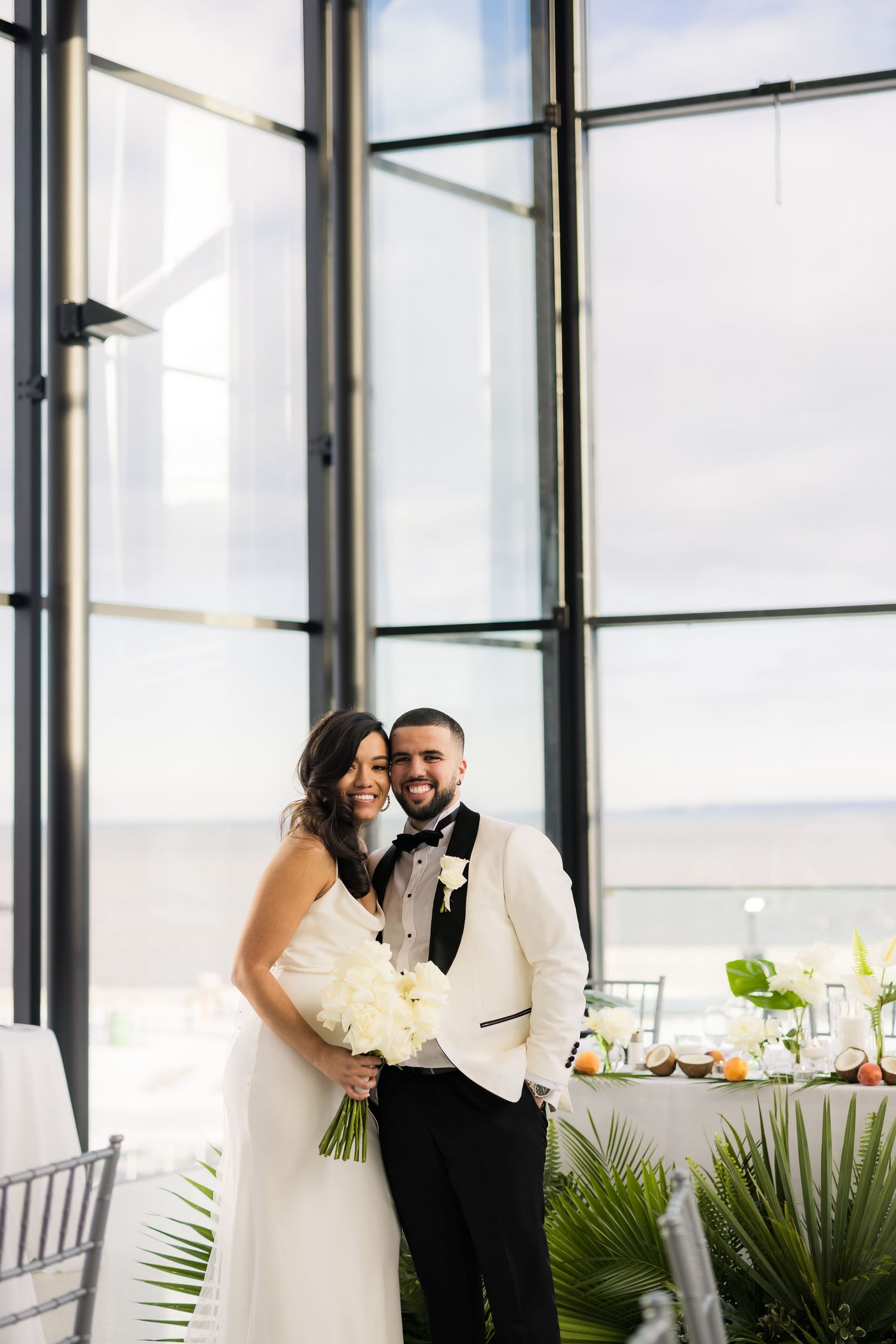 Wedding couple poses in modern venue with floor-to-ceiling windows and elegant table settings.