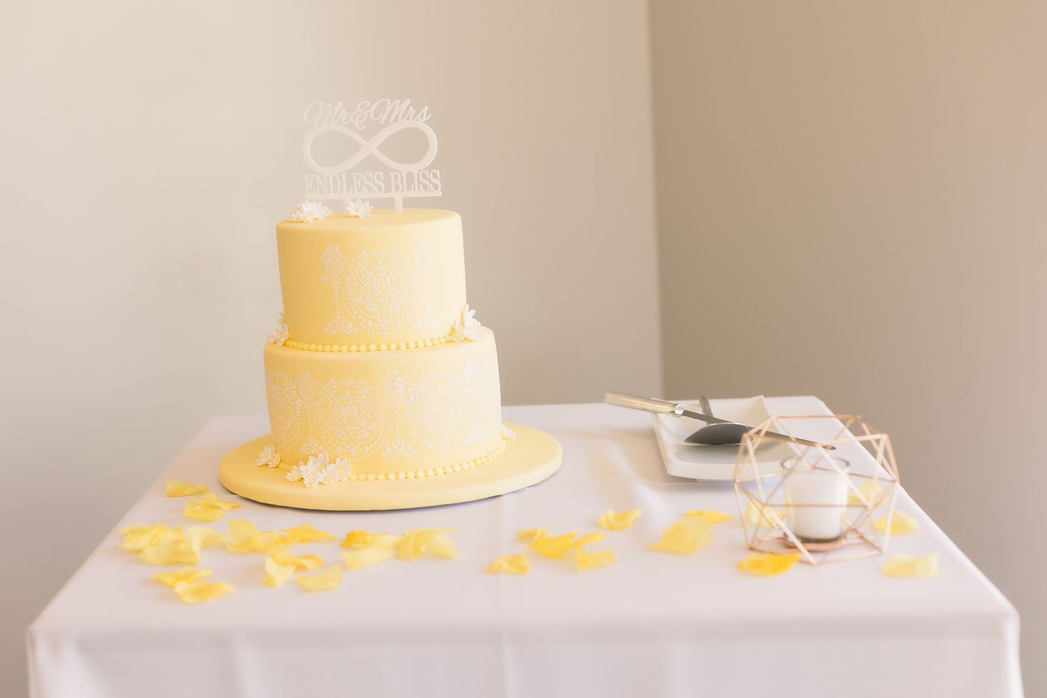 Simple two-tier yellow wedding cake with infinity symbol topper on white tablecloth.