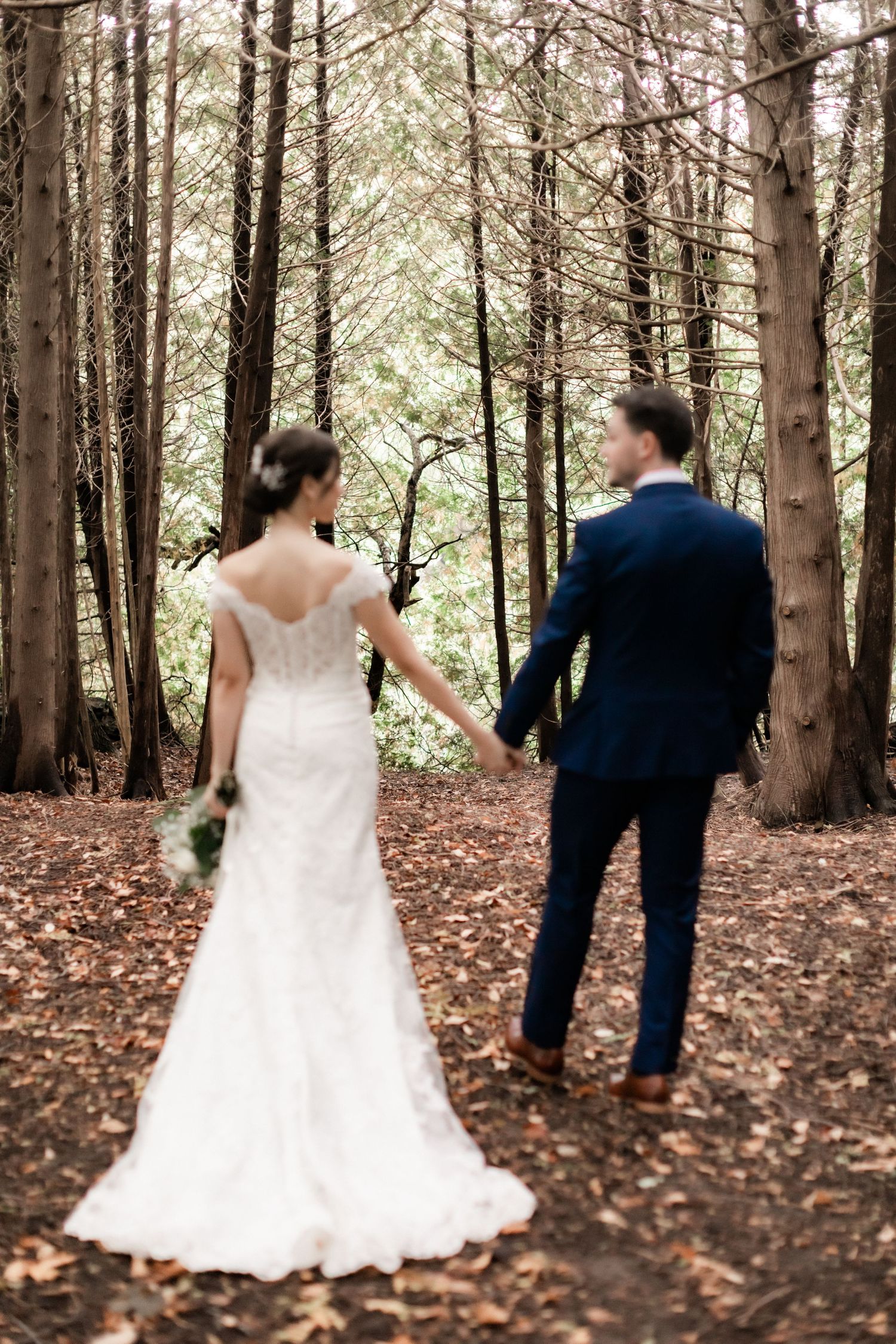Bride in lace gown and groom walking hand in hand through serene forest path.