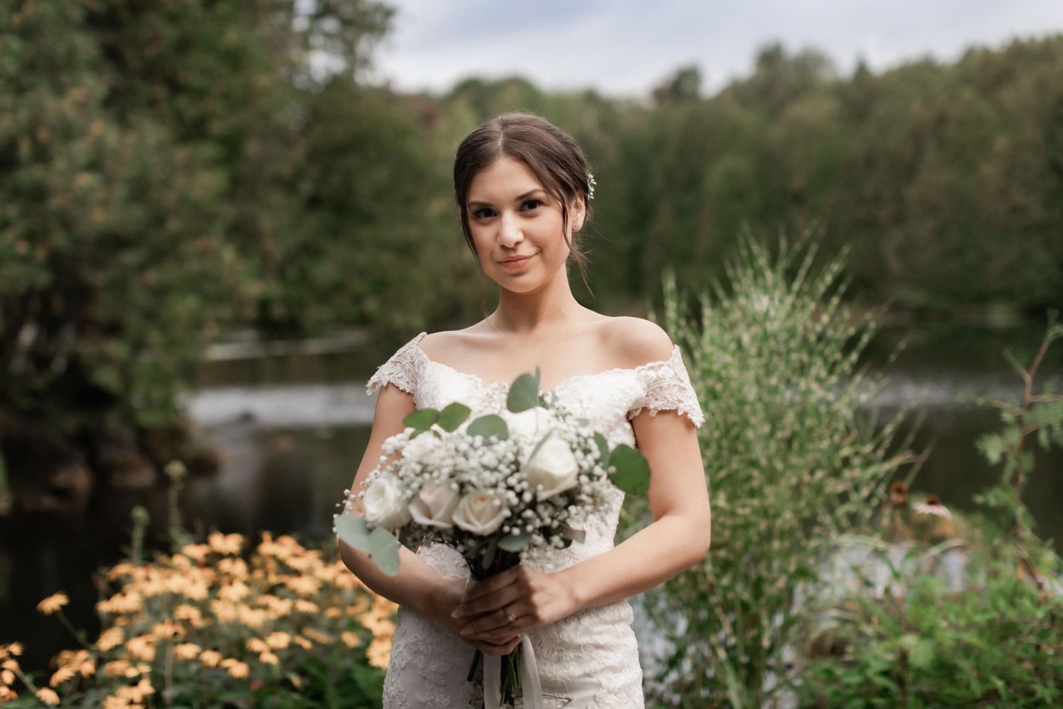 A bride in an off-shoulder lace gown holds a white bouquet in a garden setting.