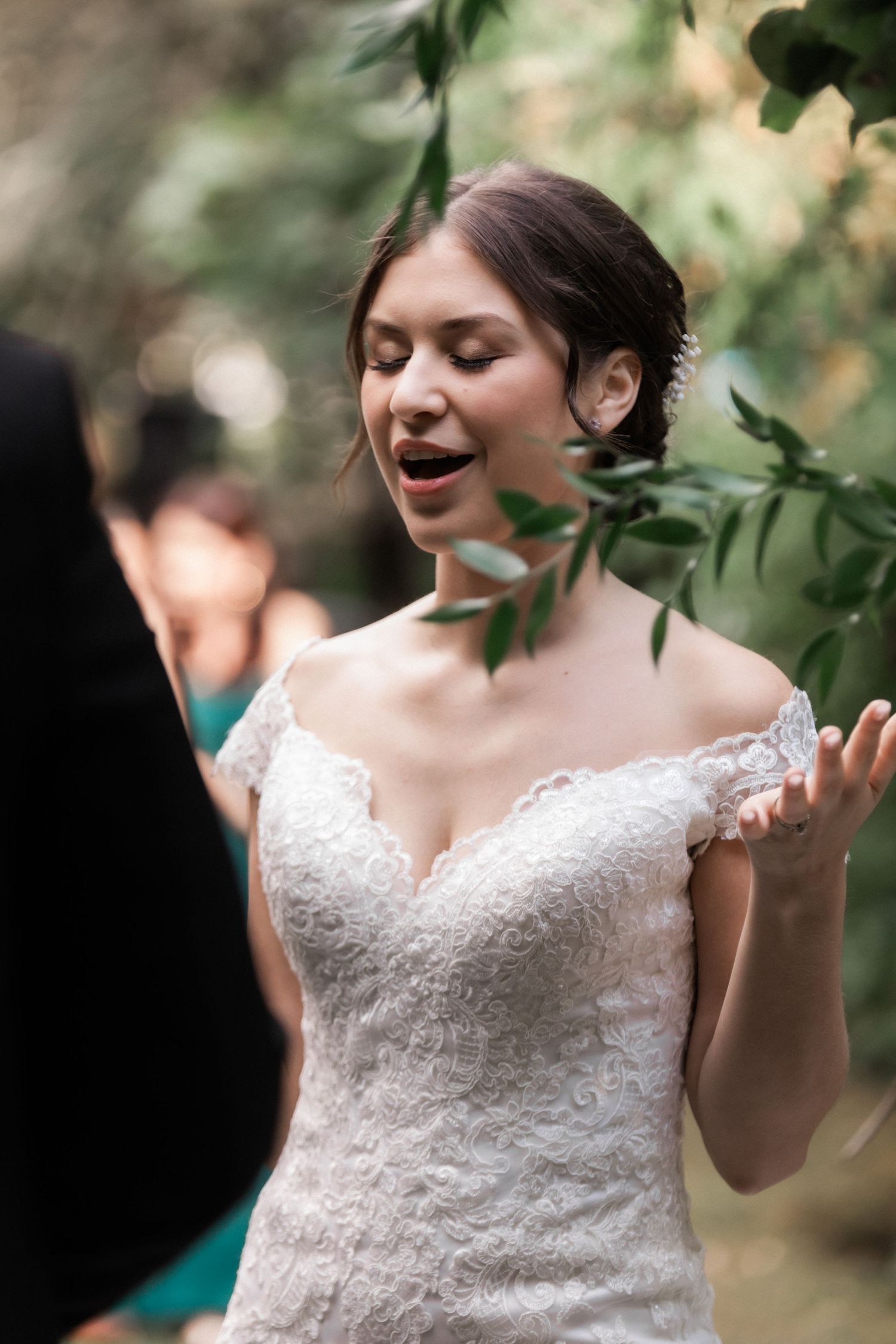 A bride in lace gown surrounded by greenery expresses joy during wedding ceremony.