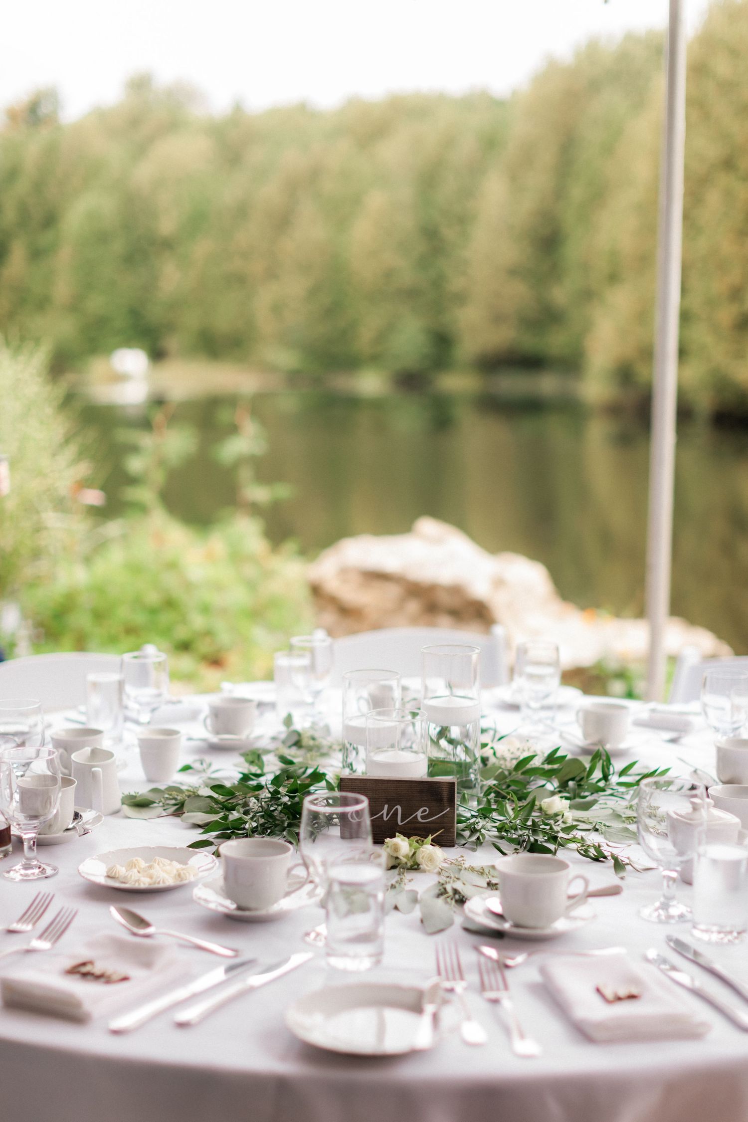 Elegant wedding table setting with white linens, greenery centerpieces and place settings by a lakeside venue.