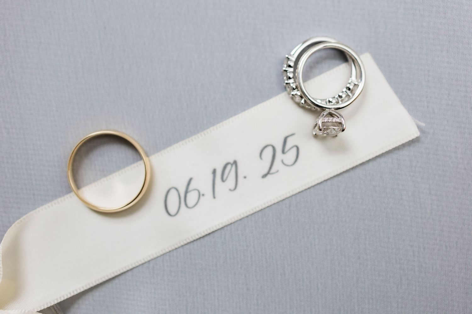 Wedding rings and a date written as '06.19.25' on a white paper strip against a gray background.