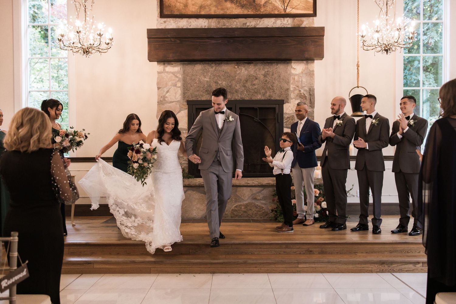 Wedding ceremony in an elegant venue with stone fireplace, chandeliers, and guests lining the aisle.