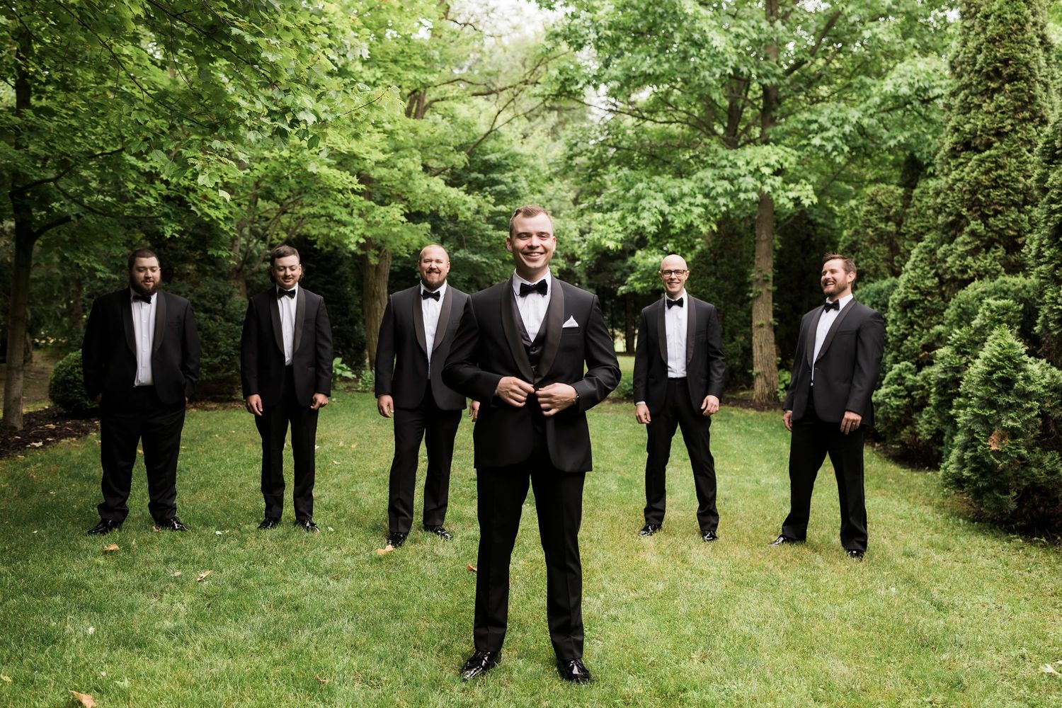 Groomsmen in black tuxedos pose together on a green lawn during an outdoor wedding.