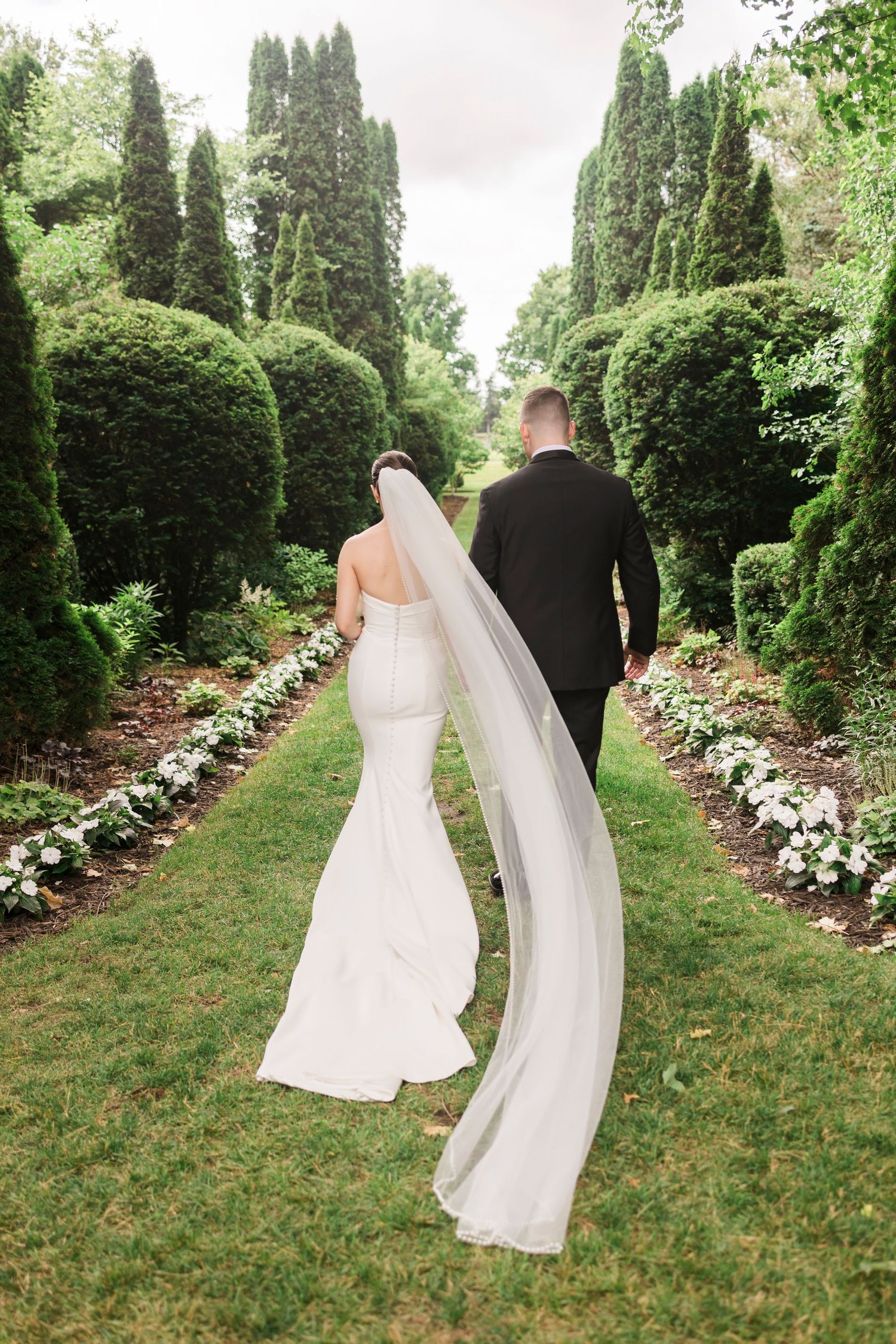Wedding couple walking through a garden path lined with tall cypress trees and white flowers.