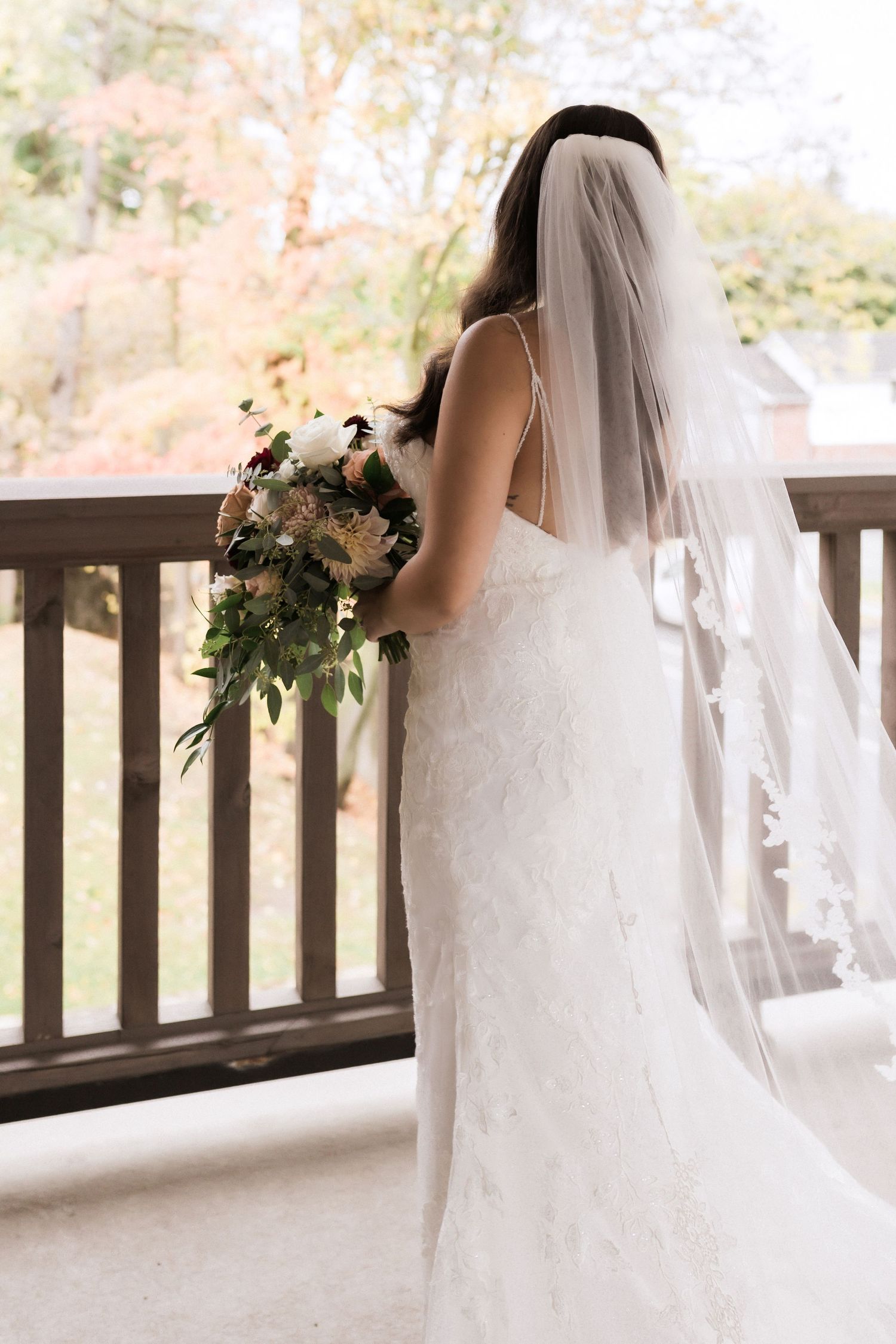 Bride in flowing white lace gown and veil stands at wooden railing holding bouquet.