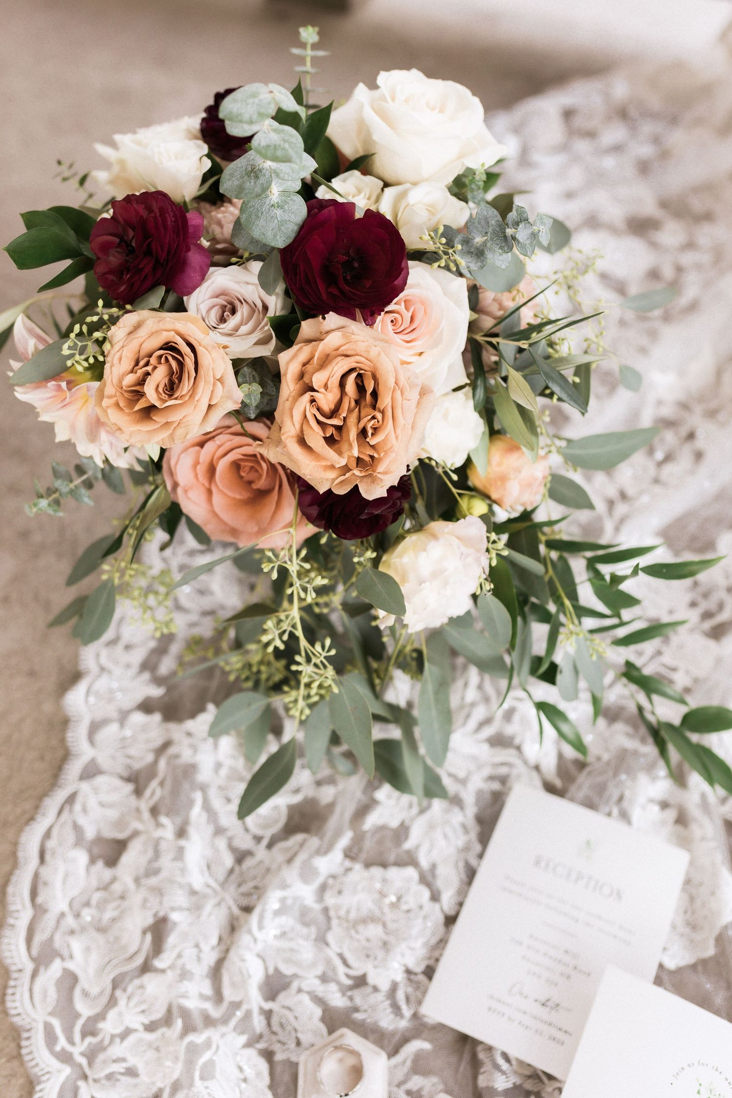 Romantic bridal bouquet with burgundy and blush roses accented with eucalyptus leaves on lace fabric.