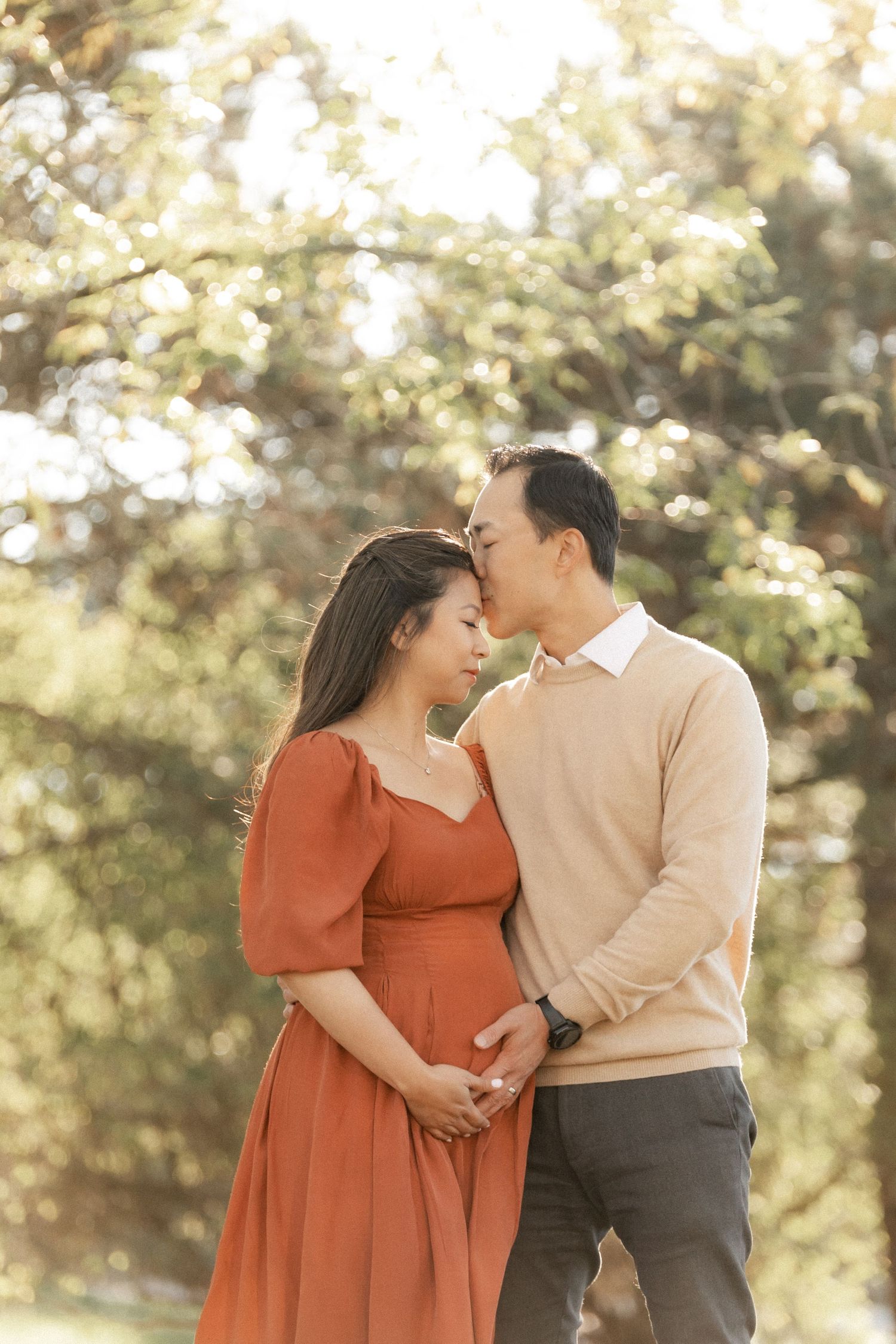 A tender maternity photoshoot in a sun-dappled forest with warm autumn lighting and coordinated outfits.