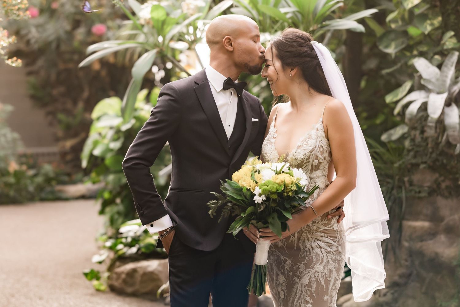 A tender wedding moment between newlyweds in a lush garden setting with tropical plants and delicate floral bouquet.