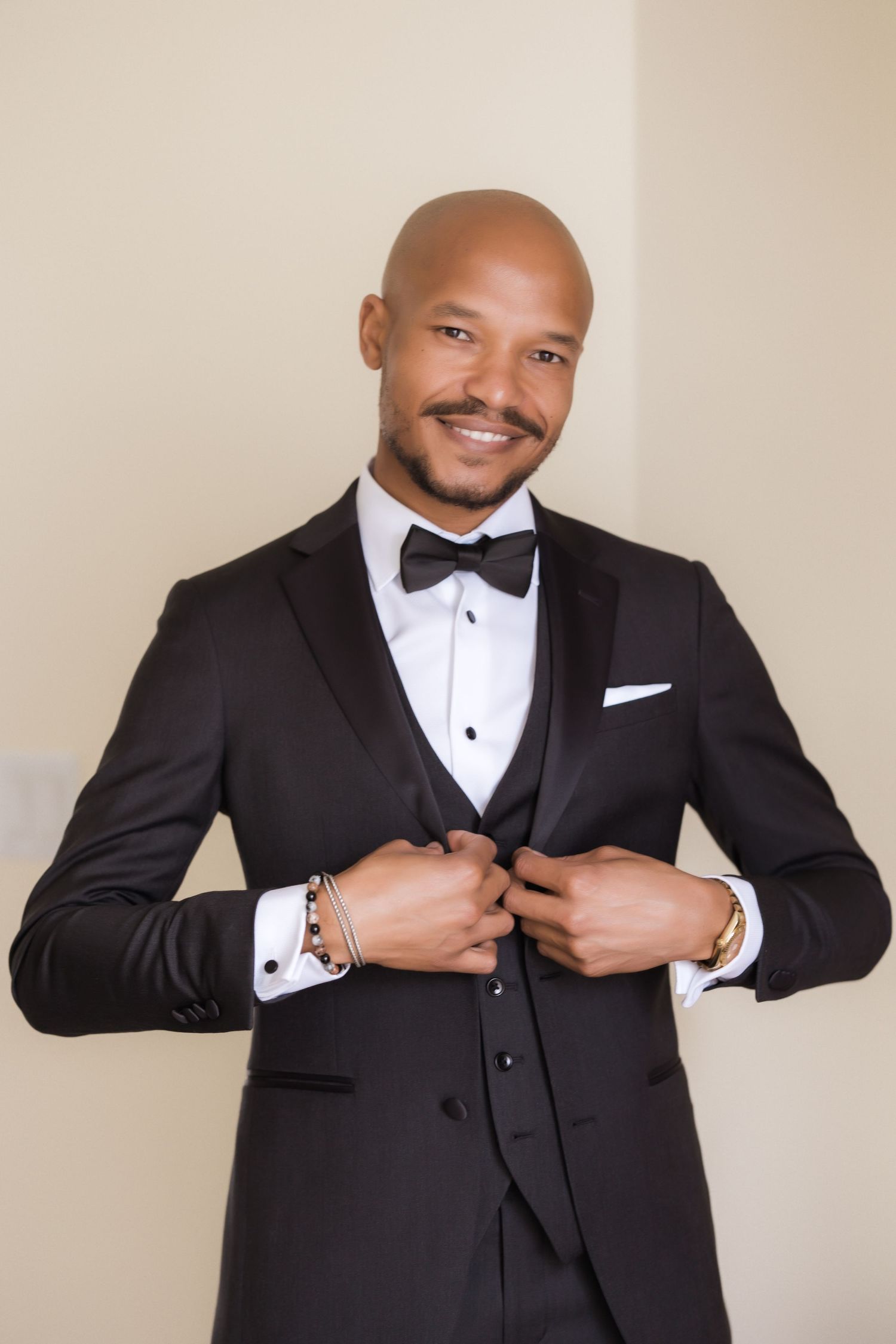 Well-dressed groom adjusts suit jacket while posing in formal black tuxedo with bow tie and three-piece styling.