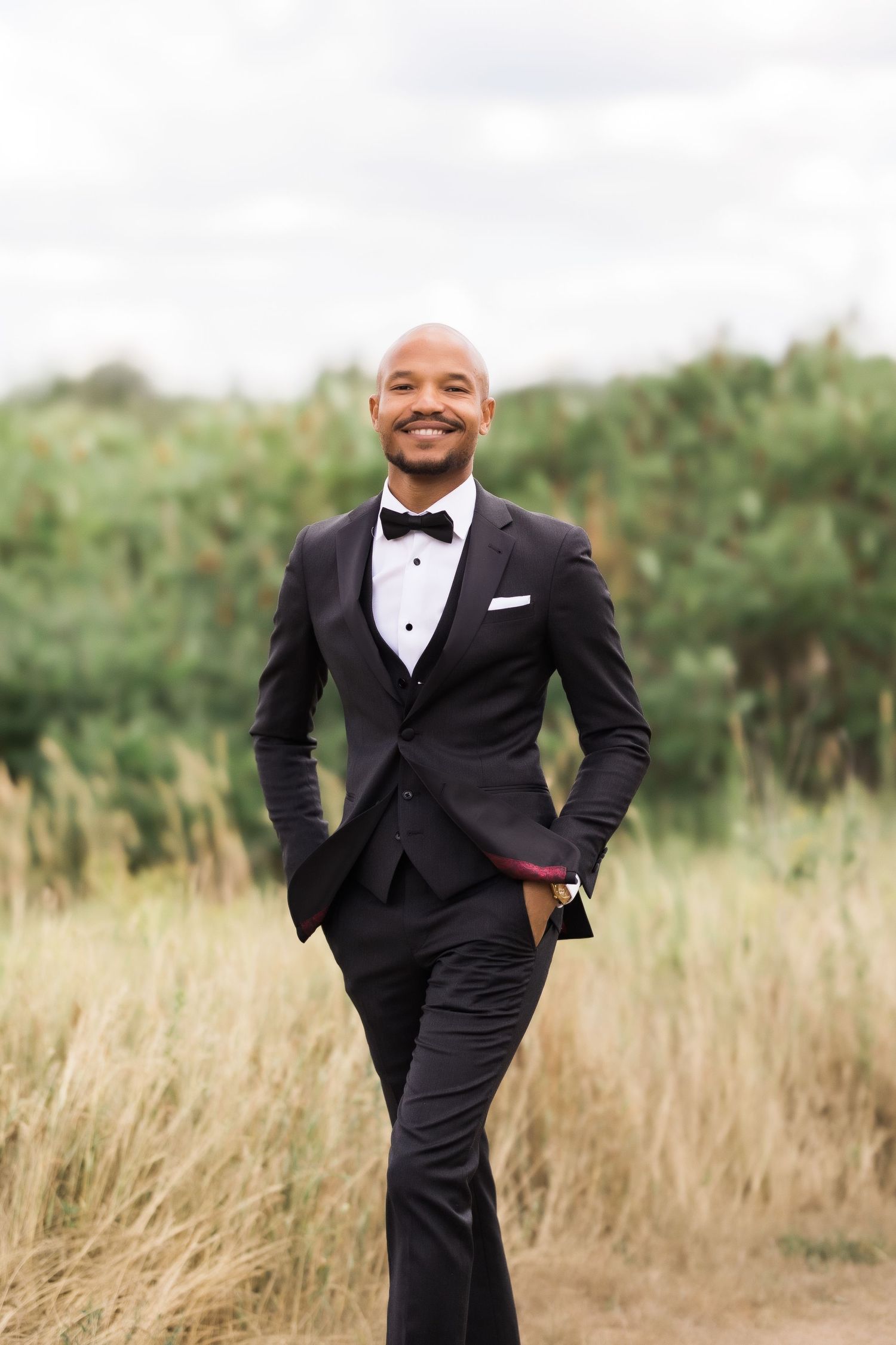 Stylish wedding portrait of person in black tuxedo standing in field of tall grass.