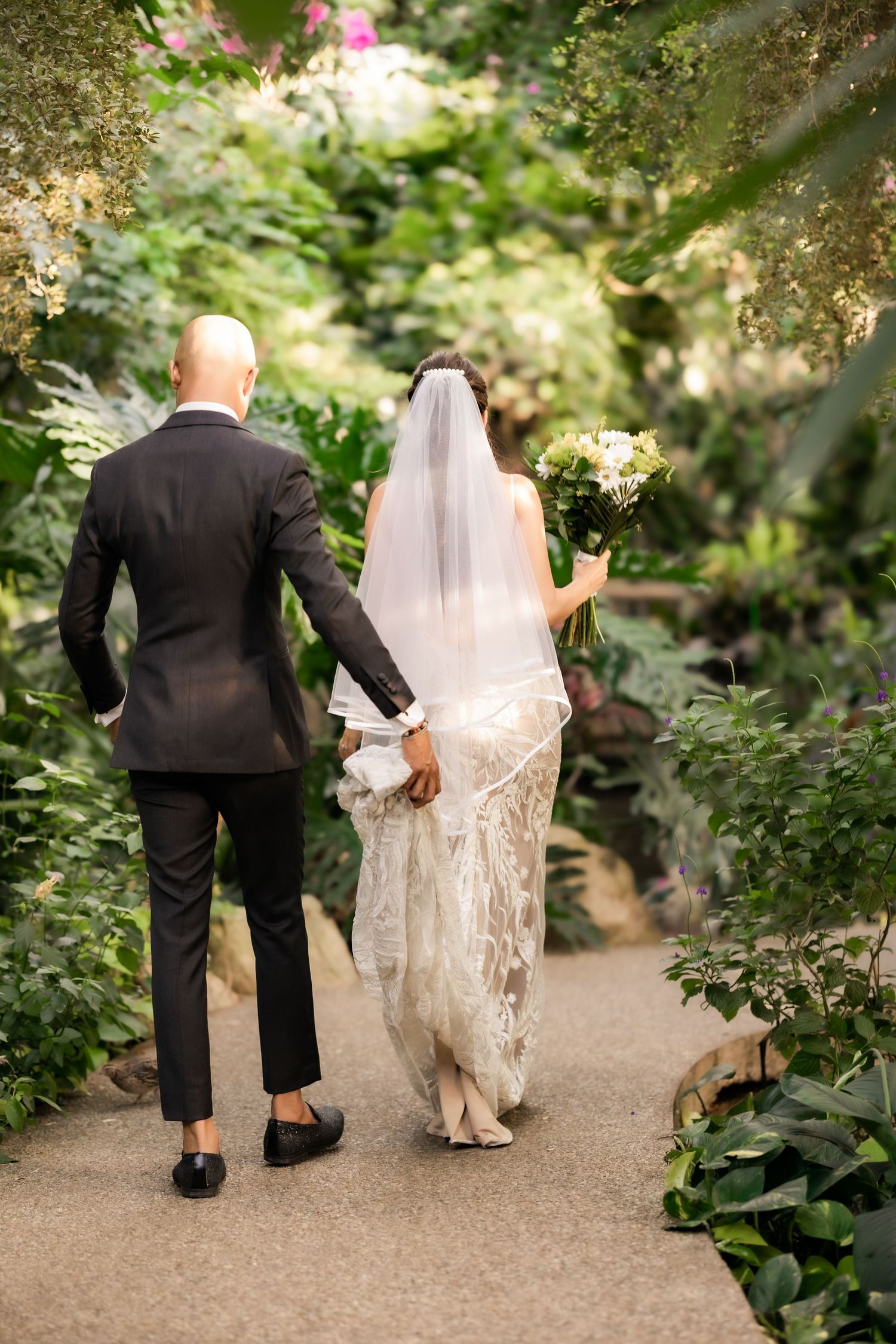 Bride and groom walking hand in hand down a garden path surrounded by lush greenery.