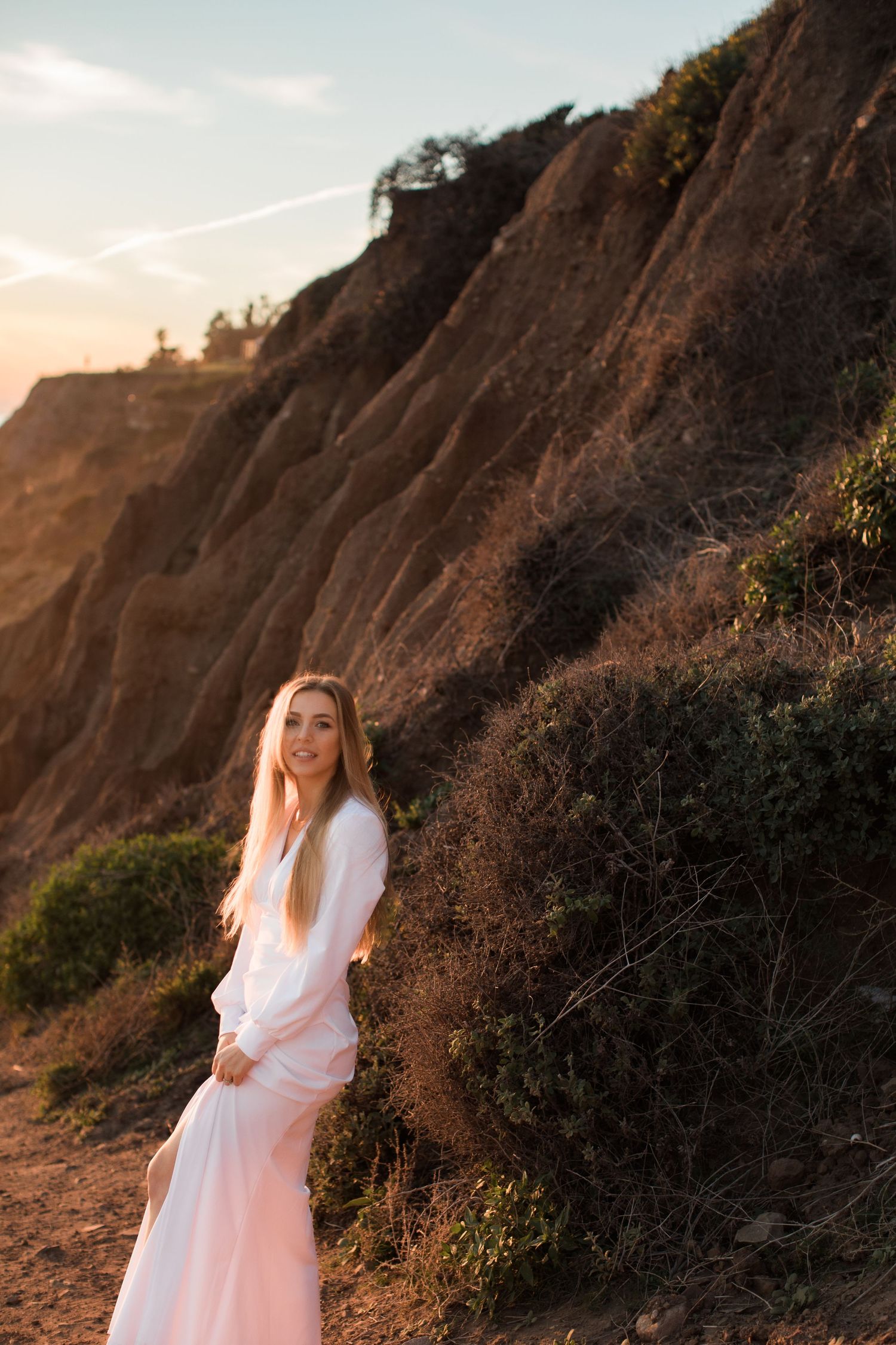 A woman in a white dress poses gracefully on a scenic coastal path at sunset.