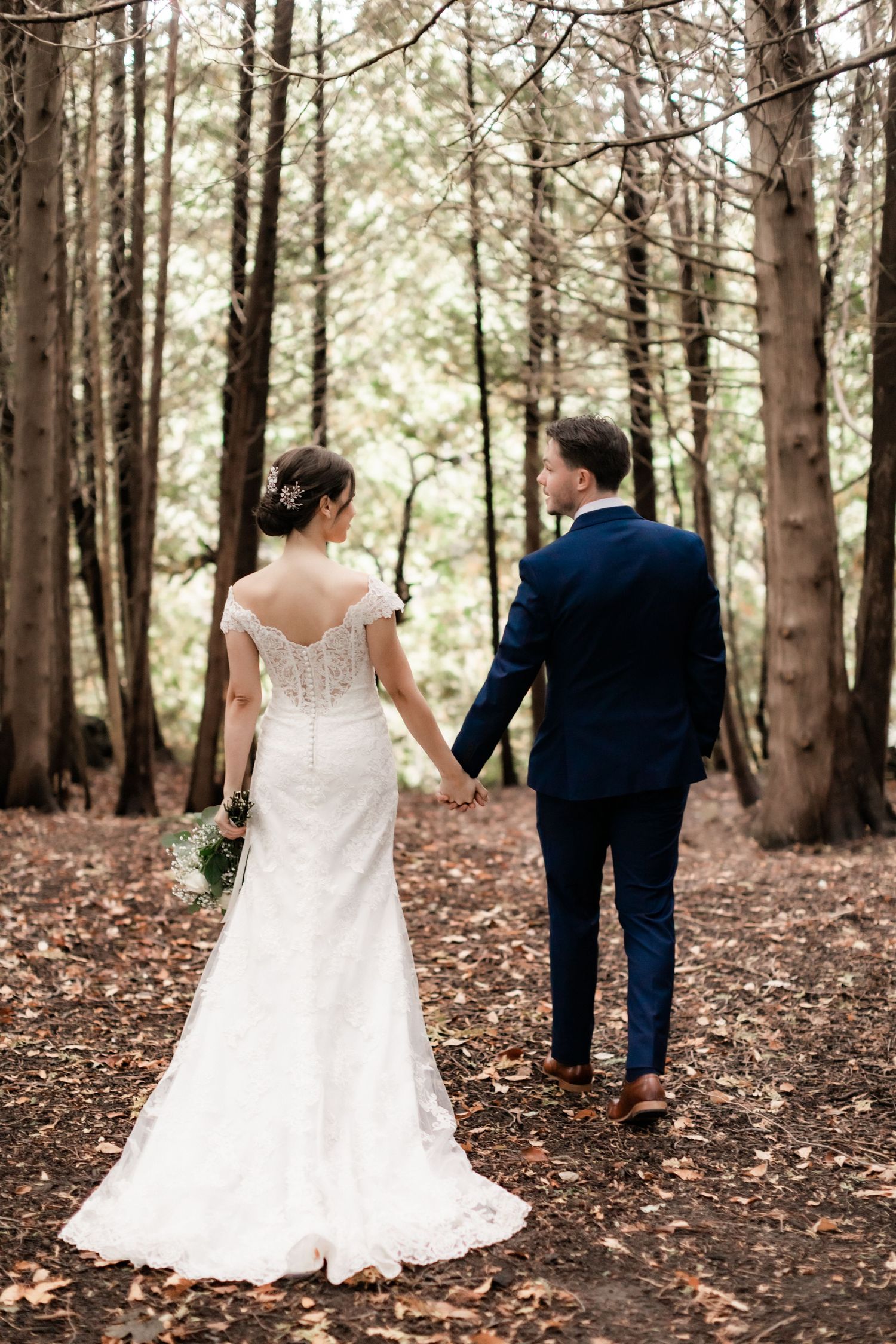 Bride and groom walking away on forest path showing detailed lace back of wedding dress.