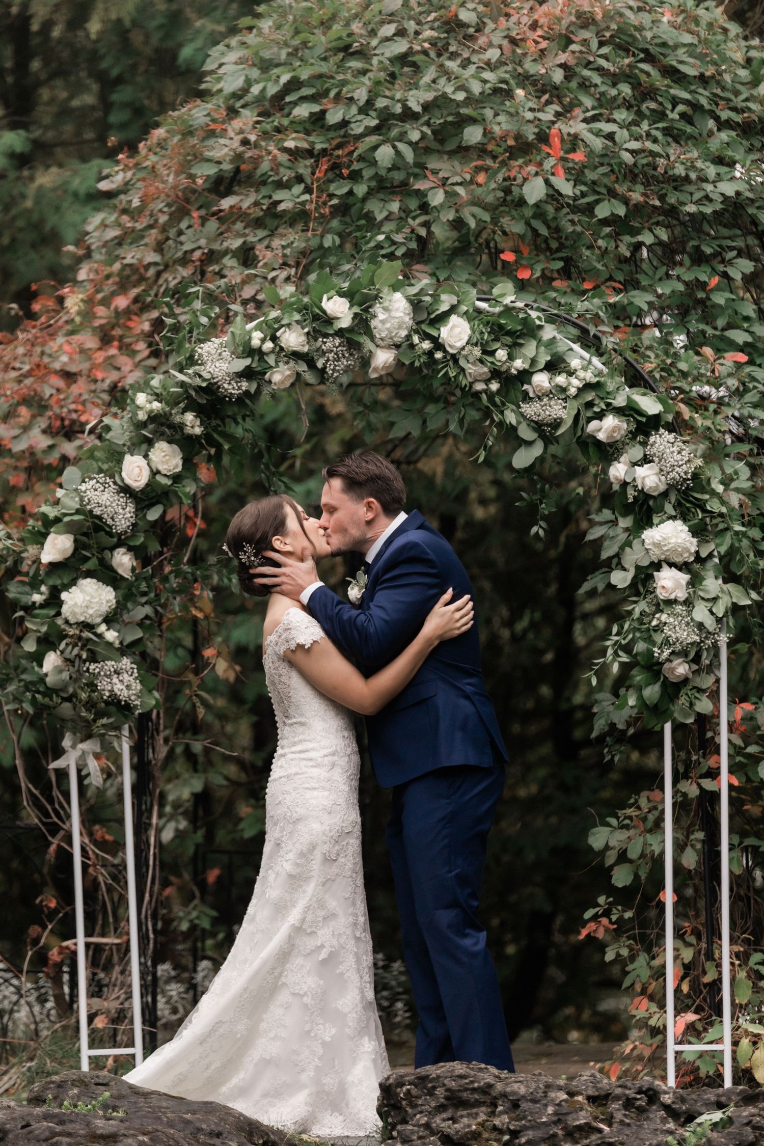 A romantic wedding moment under a floral arch decorated with white roses in a garden setting.