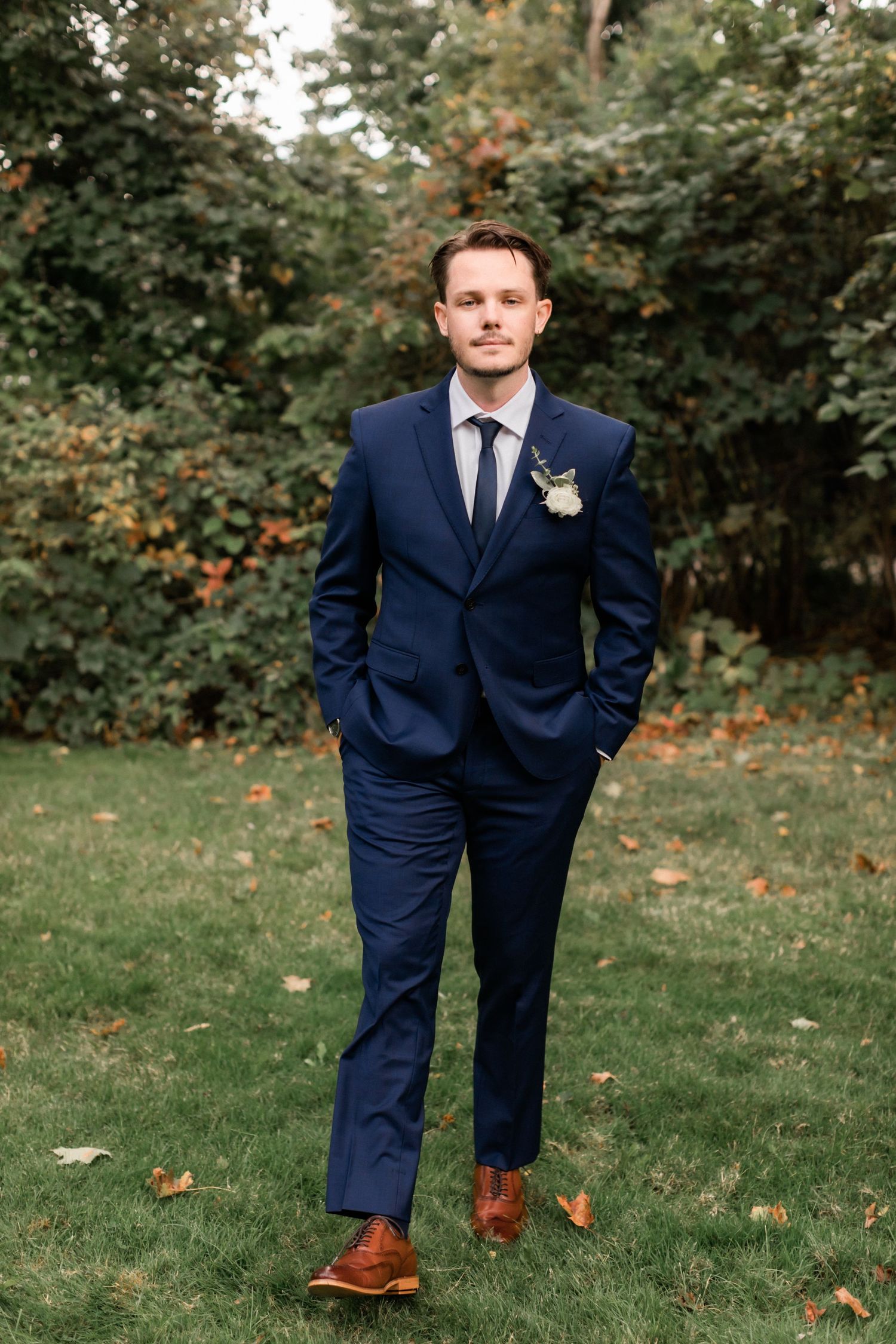 Full-length portrait of groom in navy suit and brown shoes standing on lawn.