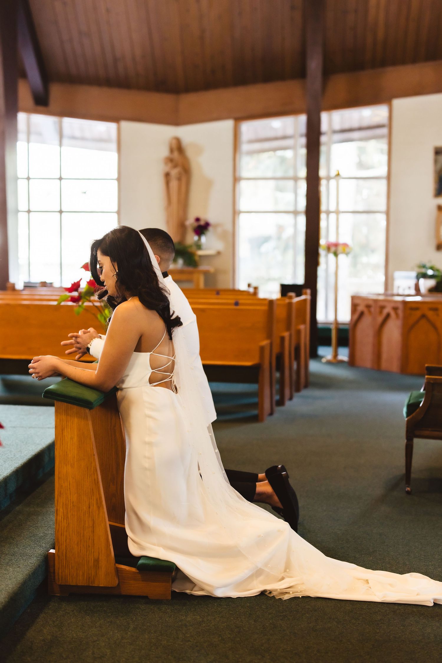 Bride praying alone in church pew wearing elegant white wedding gown with train.
