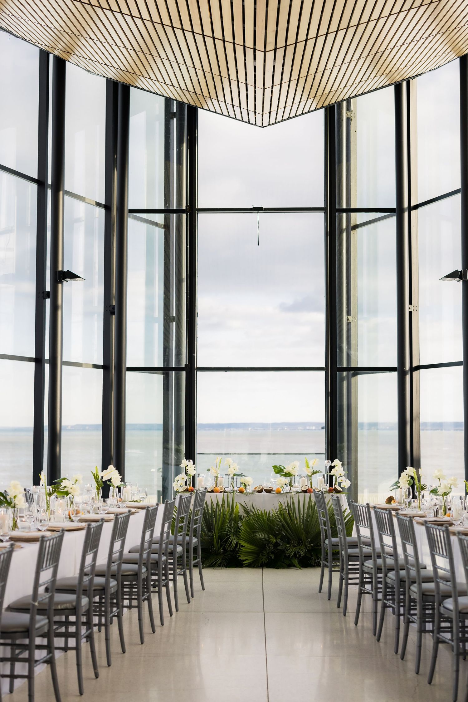 Elegant wedding reception venue with floor-to-ceiling windows overlooking water and modern table settings.
