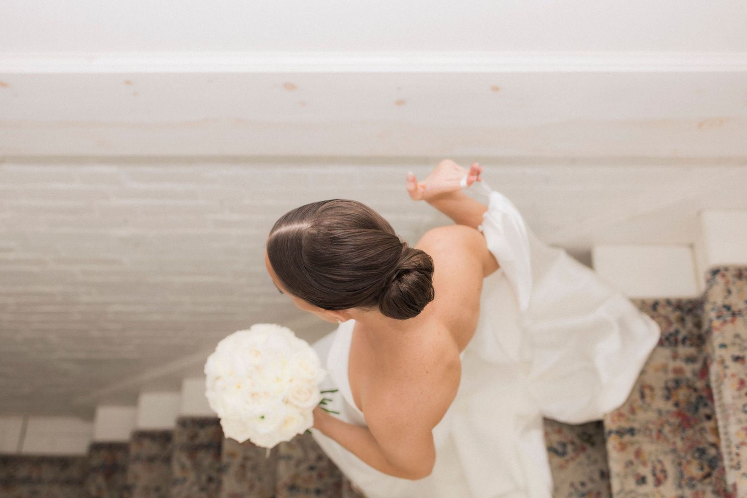 A bride in a flowing white gown stands gracefully by a stone wall in romantic sunlit outdoor setting.