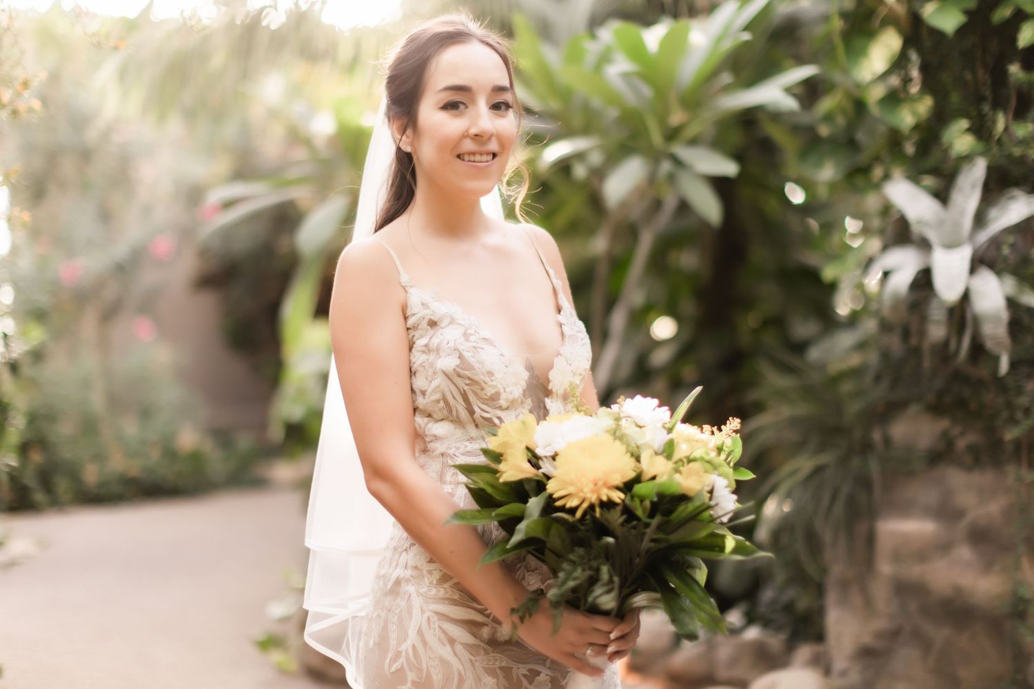 A bride in a lace wedding dress holds a bouquet of yellow roses in a lush garden setting.