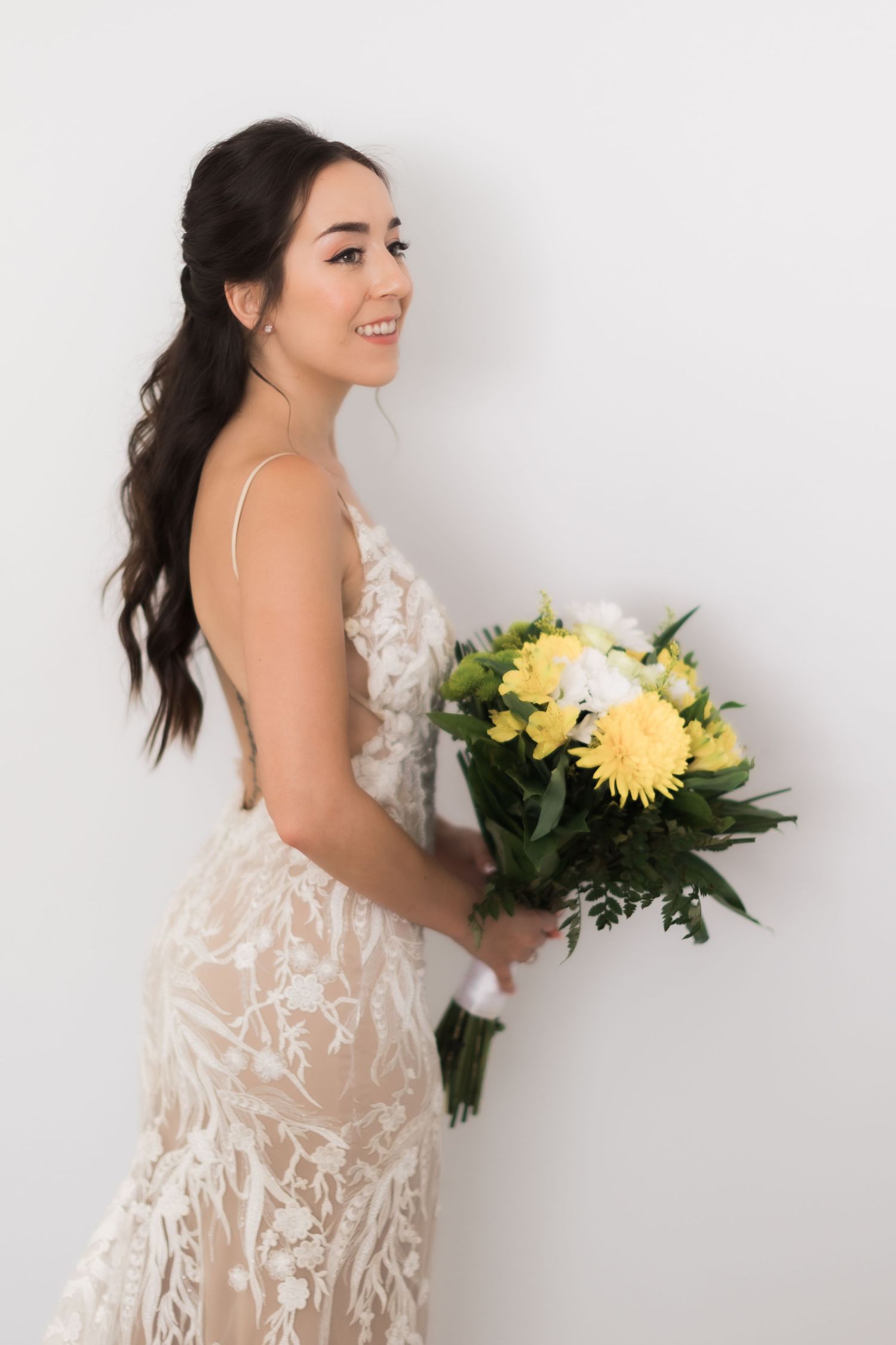 A bride in a form-fitting lace dress holds a yellow and white bouquet against a white backdrop.